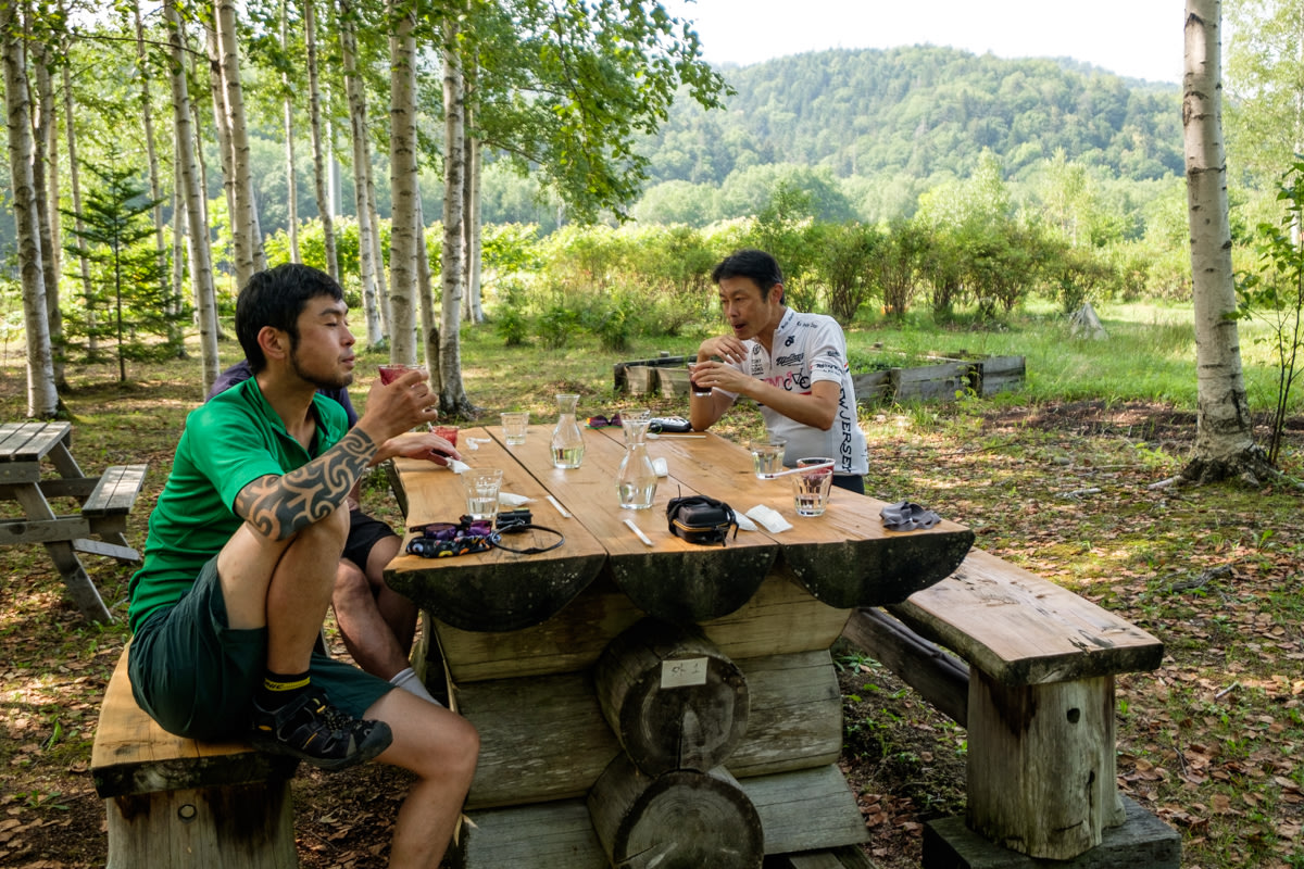 Two cyclists relax drinking berry juice while sat on a bench under Japanese White Birch trees.