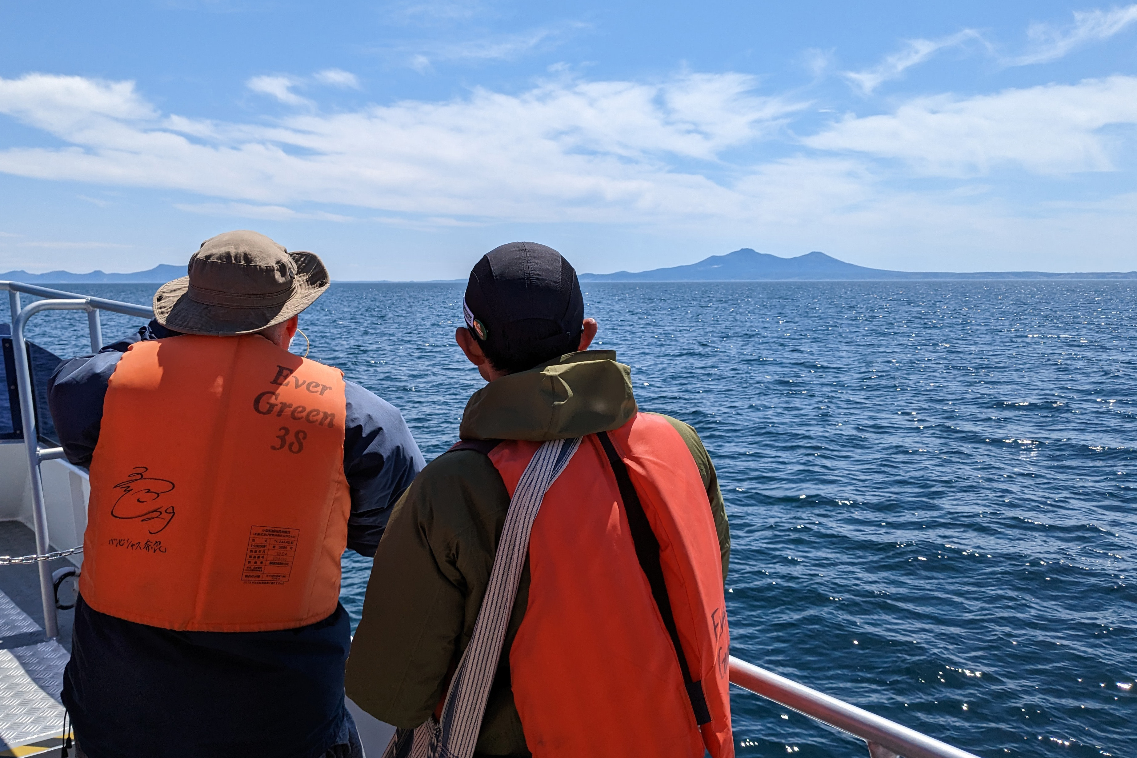 Two people scan the ocean for whales off the coast of the Shiretoko peninsula. There a a few white clouds in the sky and a mountain is seen in the distance.