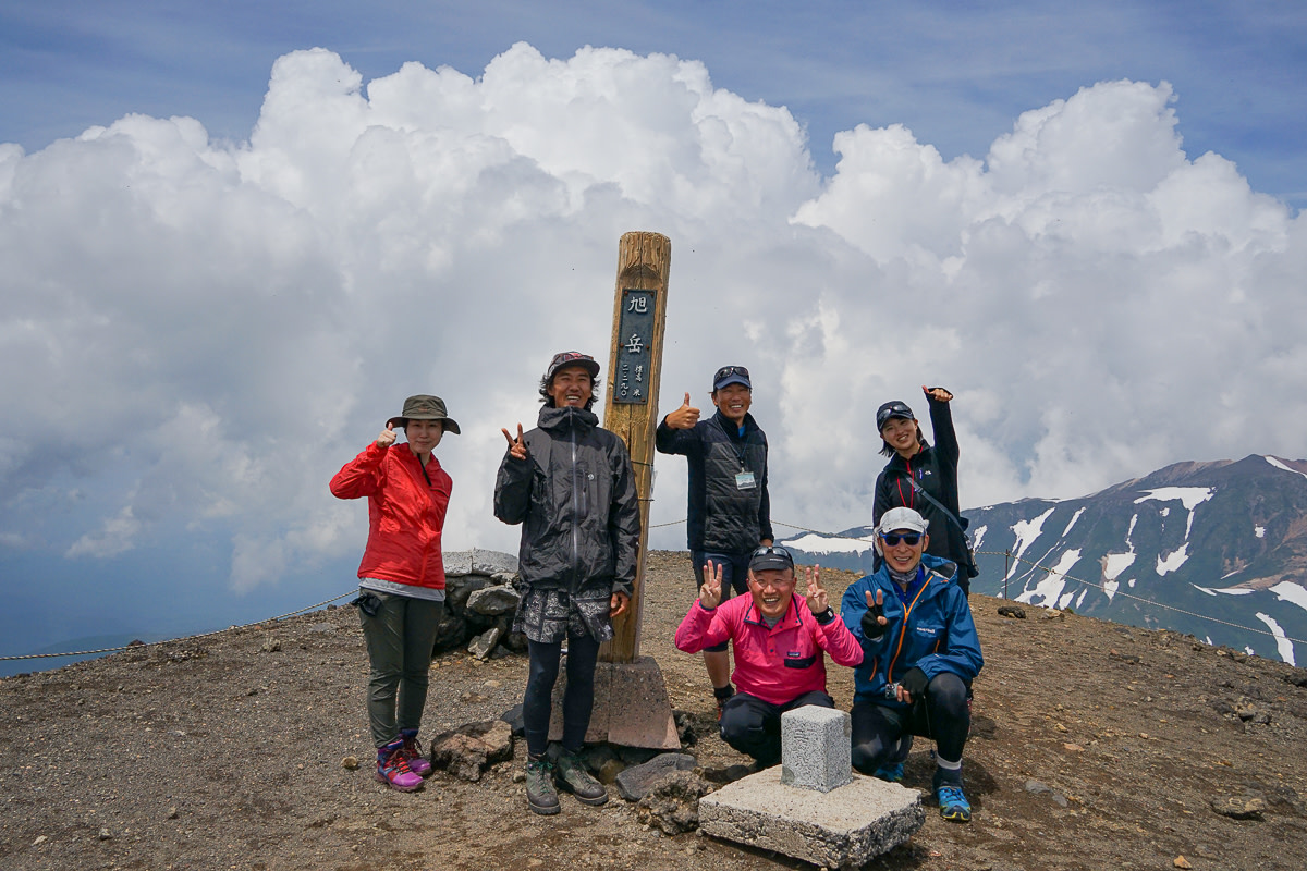 A group of hikers smile and pose for the camera. They are standing around a post marking the summit of Mt Asahidake. A fluffy white cloud fills the sky behind them.