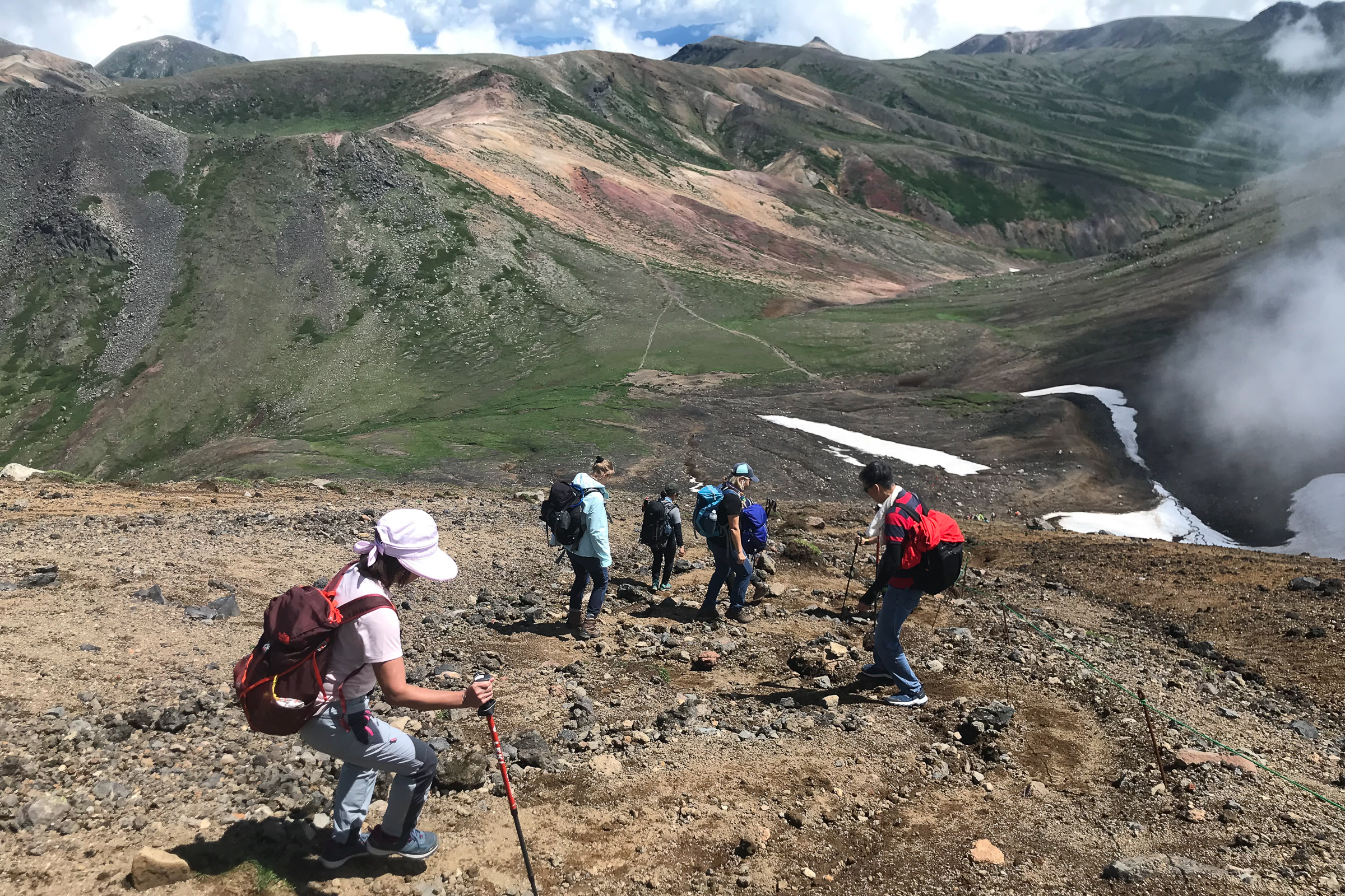 A group of hikers descend a rocky mountain slope. It is a sunny day and there are lingering snows around.