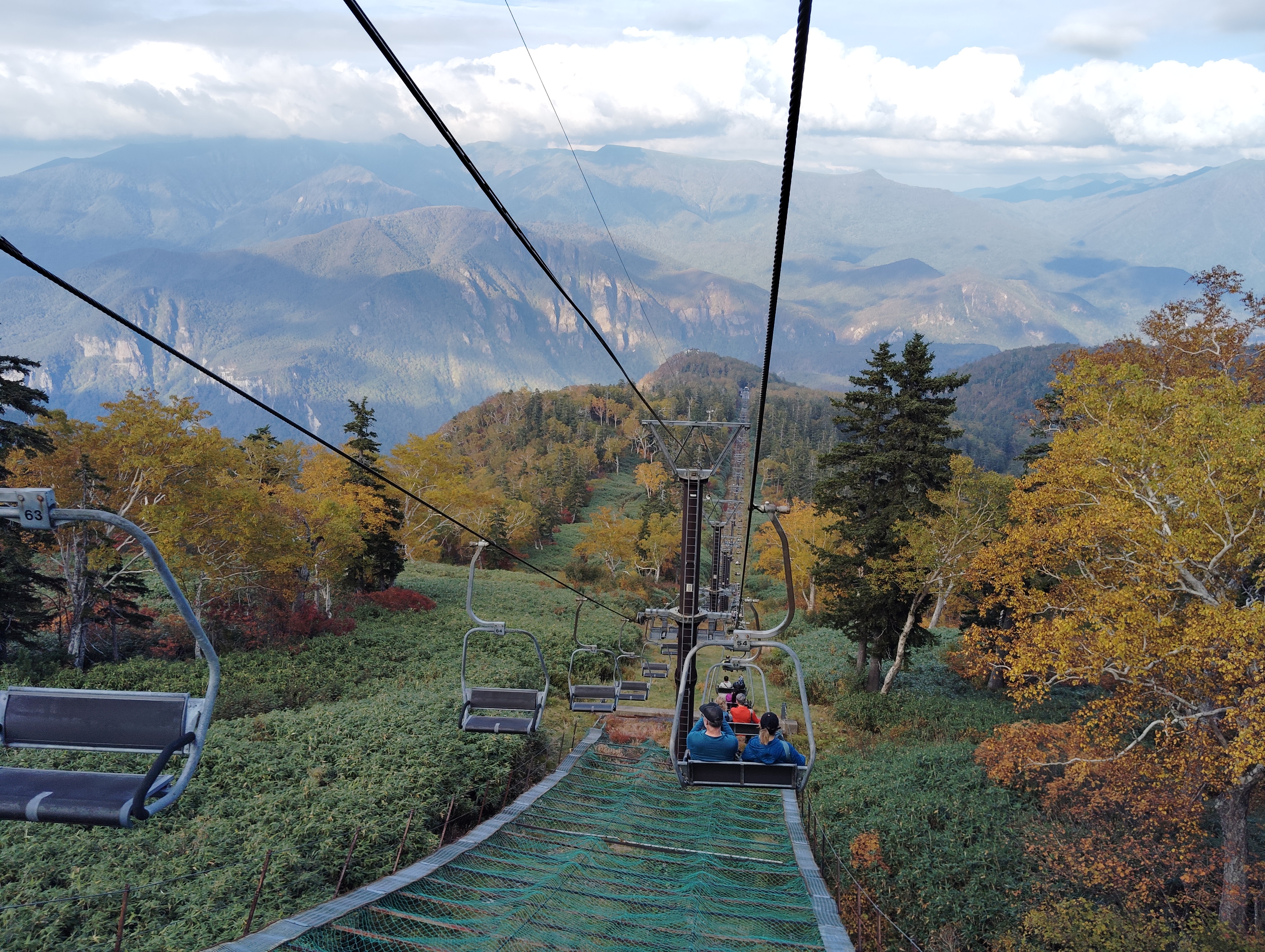 A photo taken from behind of a group of people descending a mountain in a chair lift. There are two people sat in the chair.