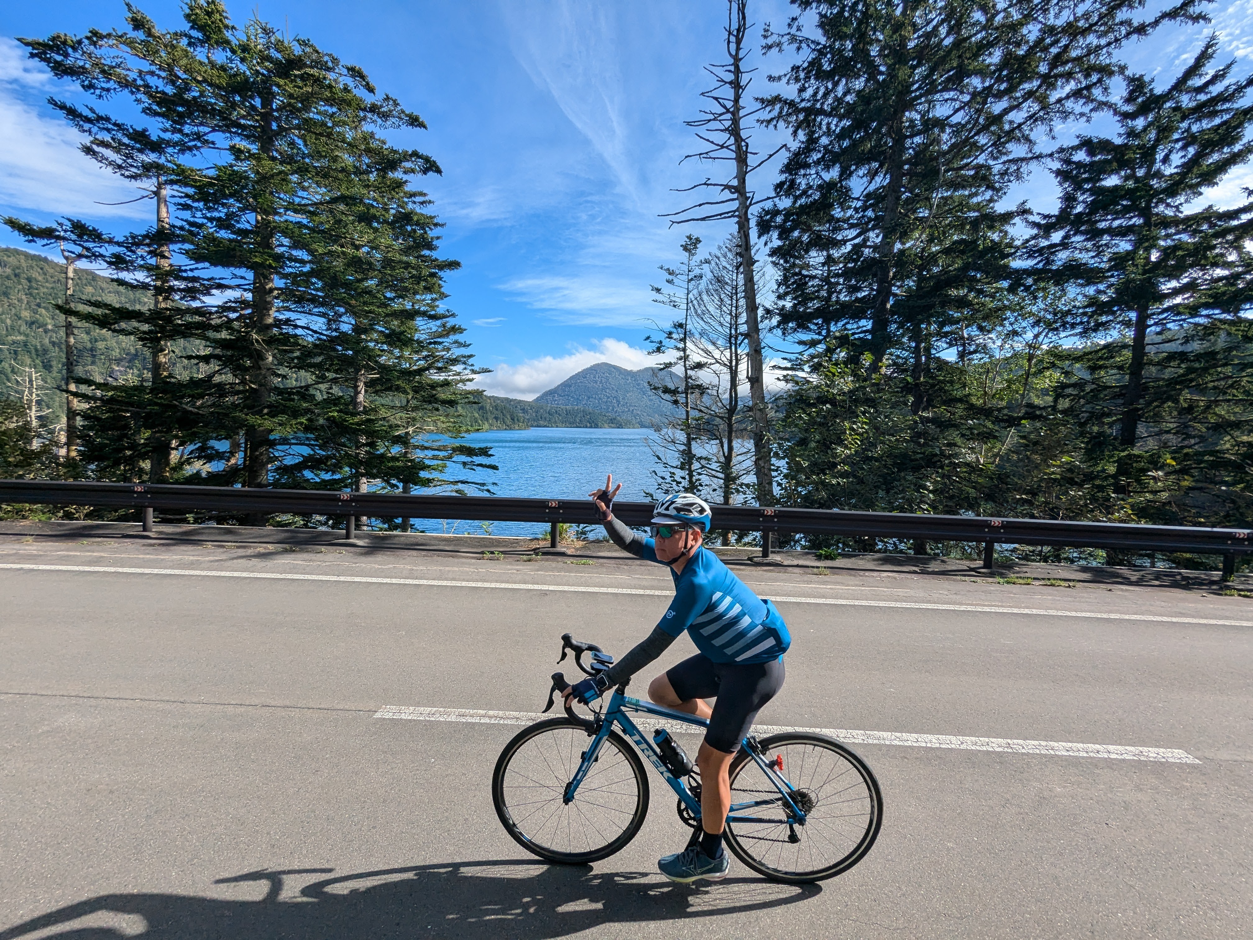 A cyclist cycles along a road, giving a "peace" or "victory" sign to the camera. Lake Shikaribetsu is in the background. It is a beautiful, sunny day.