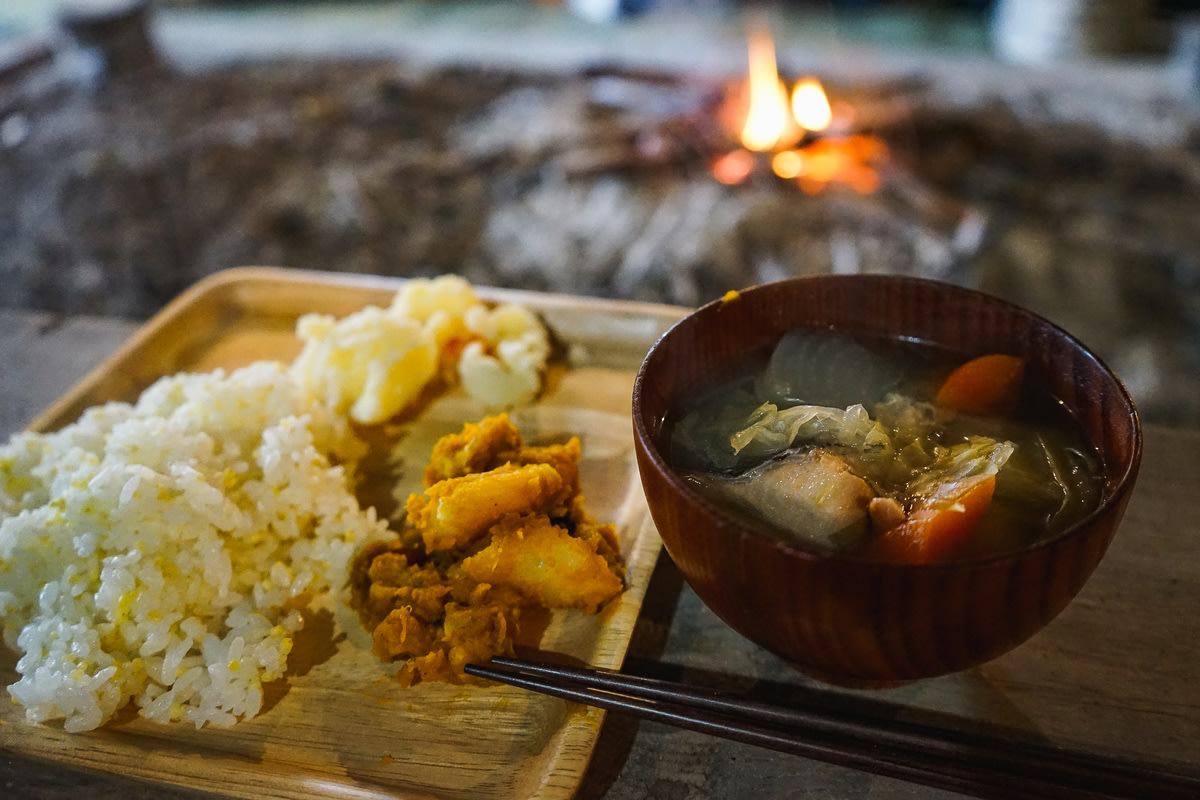 A traditional Ainu meal in front of a fire, featuring a bowl of fish broth with fish and vegetables and a millet and rice mix. Root vegetable dishes made from pumpkin and potato are also served.