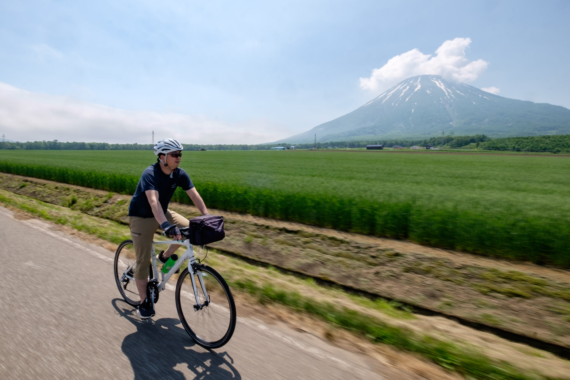 A cyclist rides past farm land with a volcano in the background.