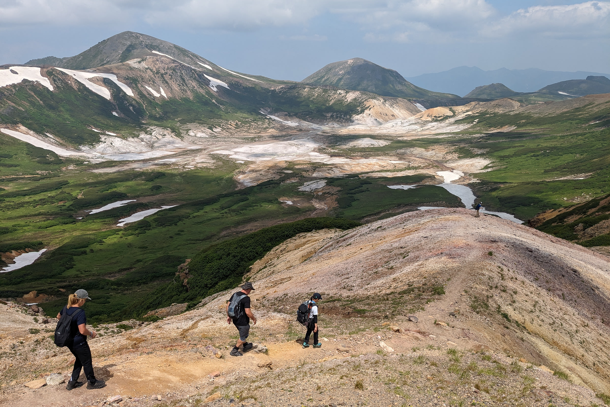 Three hikers descend down a mountain ridge line, in view of the Ohachidaira Caldera. There is lingering snow on the mountains around the caldera.