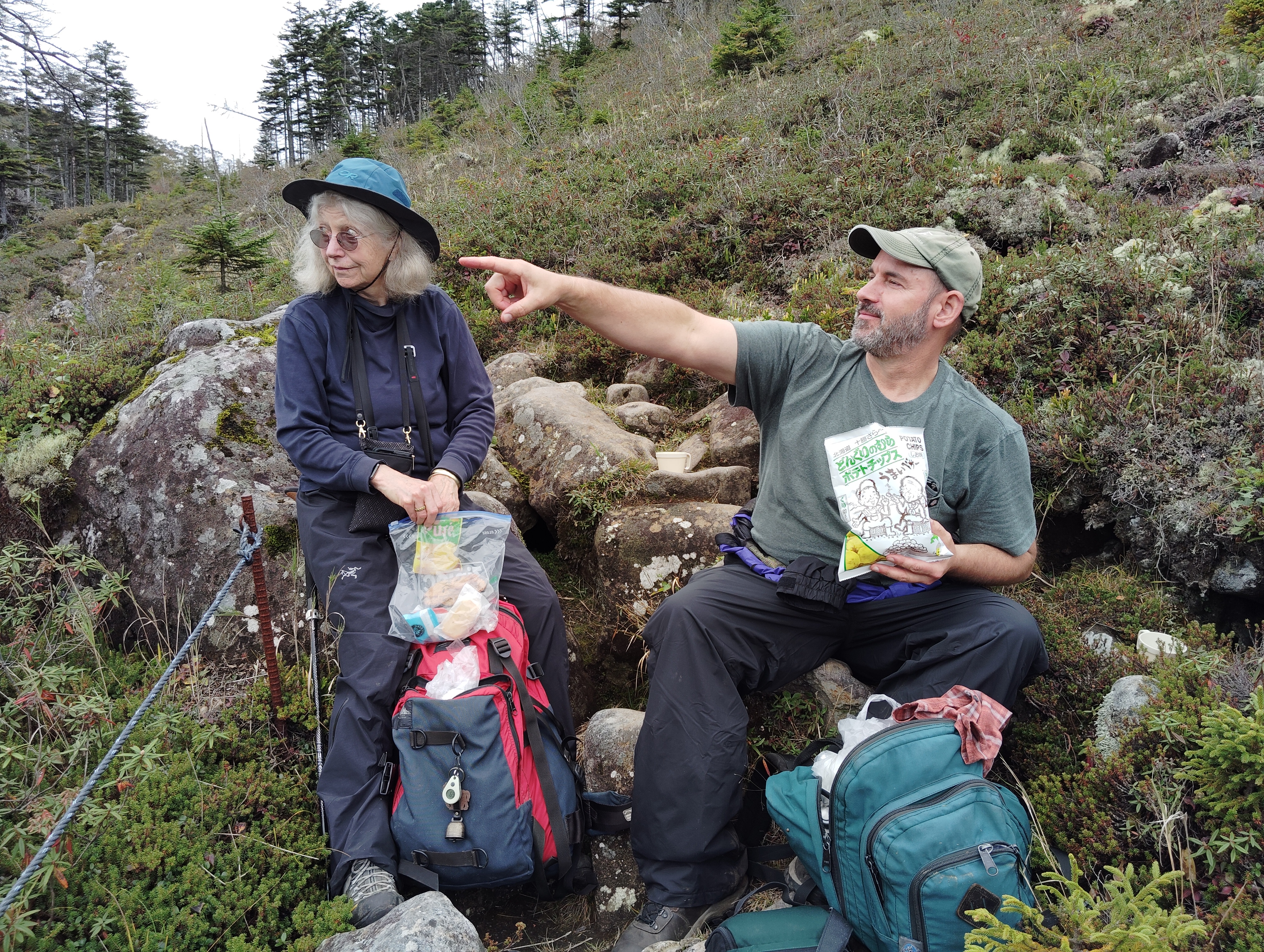 Two hikers, a man and a woman, sit on boulders at the top of a mountain for their lunch. It is an overcast day. The man holds a bag of potato chips and is pointing out towards the horizon. The woman is looking at where he is pointing.