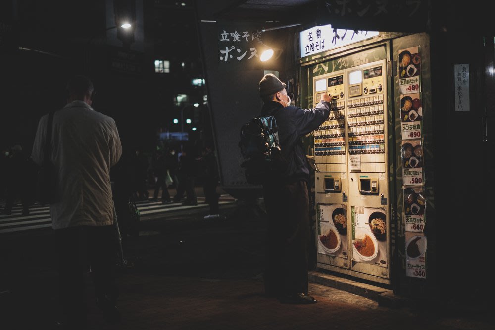 A photo at night of a man standing in front of a Japanese ticket machine for a soba noodle restaurant. The ticket machine has lots of buttons, each one for a menu item.