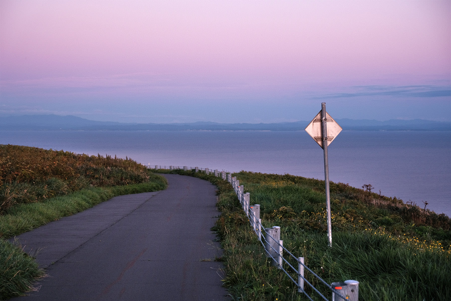 A narrow road runs along the seaside near Haboro in Hokkaido. It is evening and the sky is a pastel pink colour.