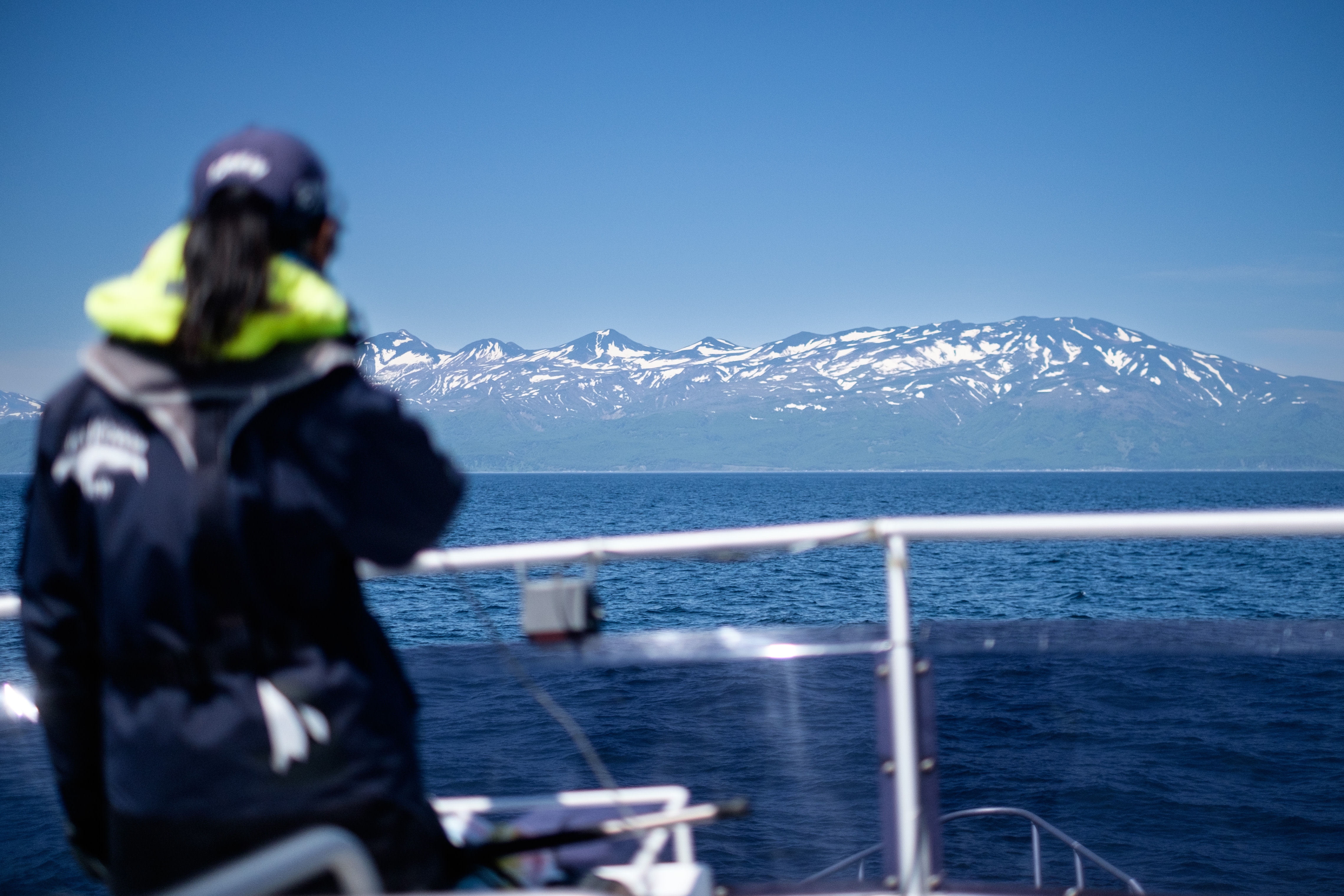 A crew member of a whale watching cruise looks out from the top deck of a boat. Across the water you can see the mountains of the Shiretoko peninsula, which still have patches of snow on them.