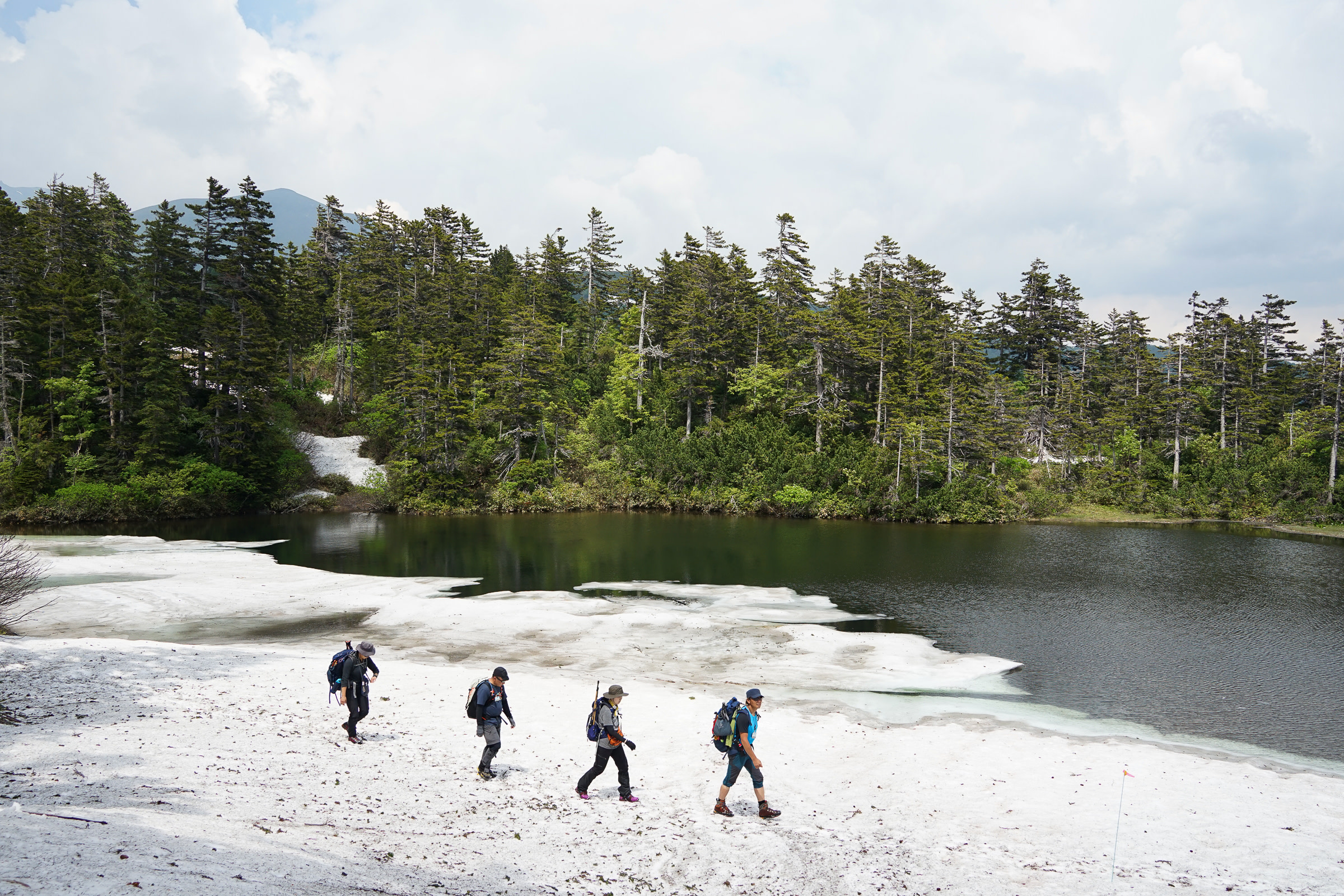 Four hikers walk upon snow beside a pond at Kogen Numa.