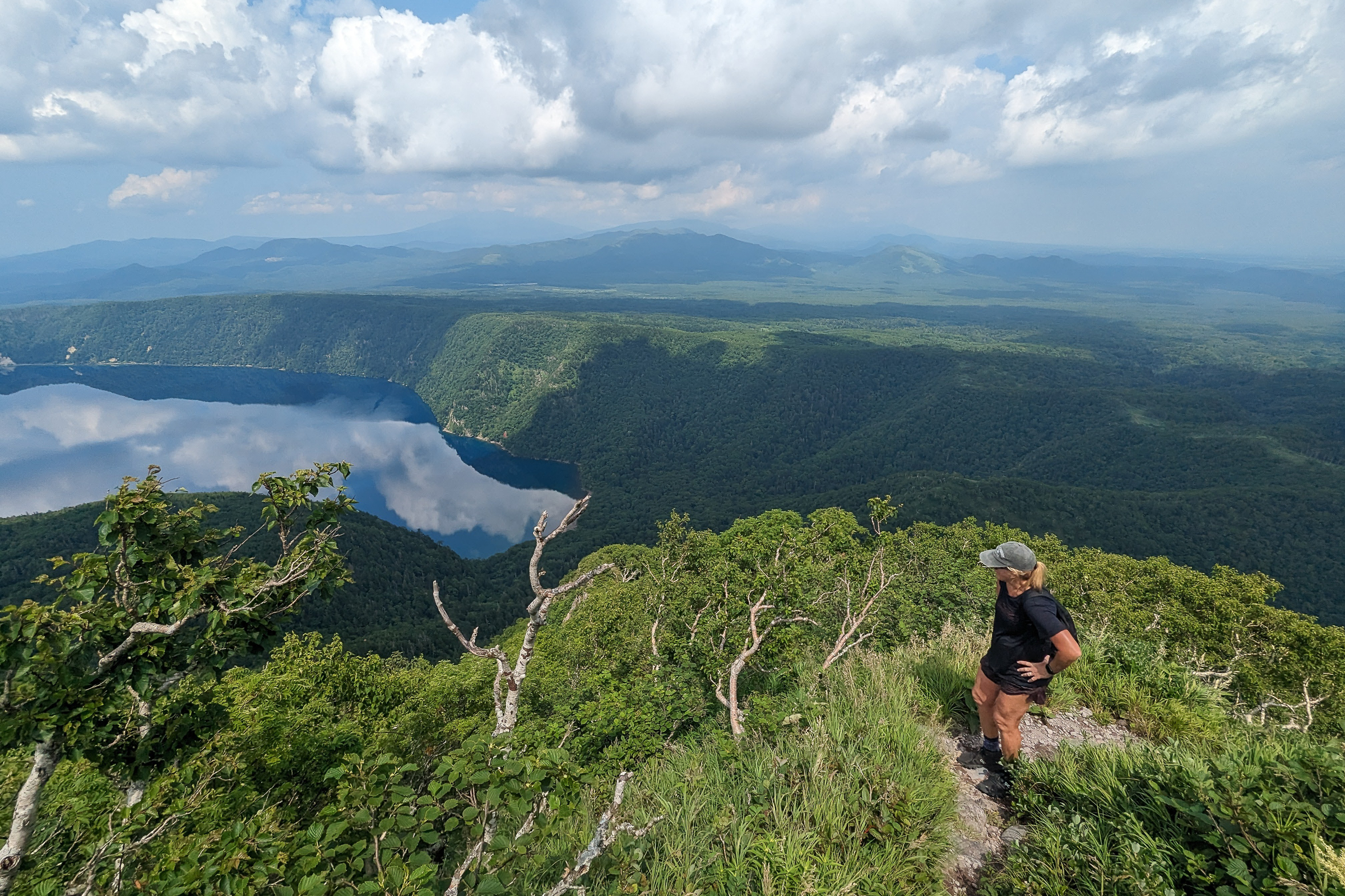 A female hiker pauses on a hike to admire the view of Lake Mashu on a lovely day. It is partially cloudy and the lake's surface is reflecting the sky. 