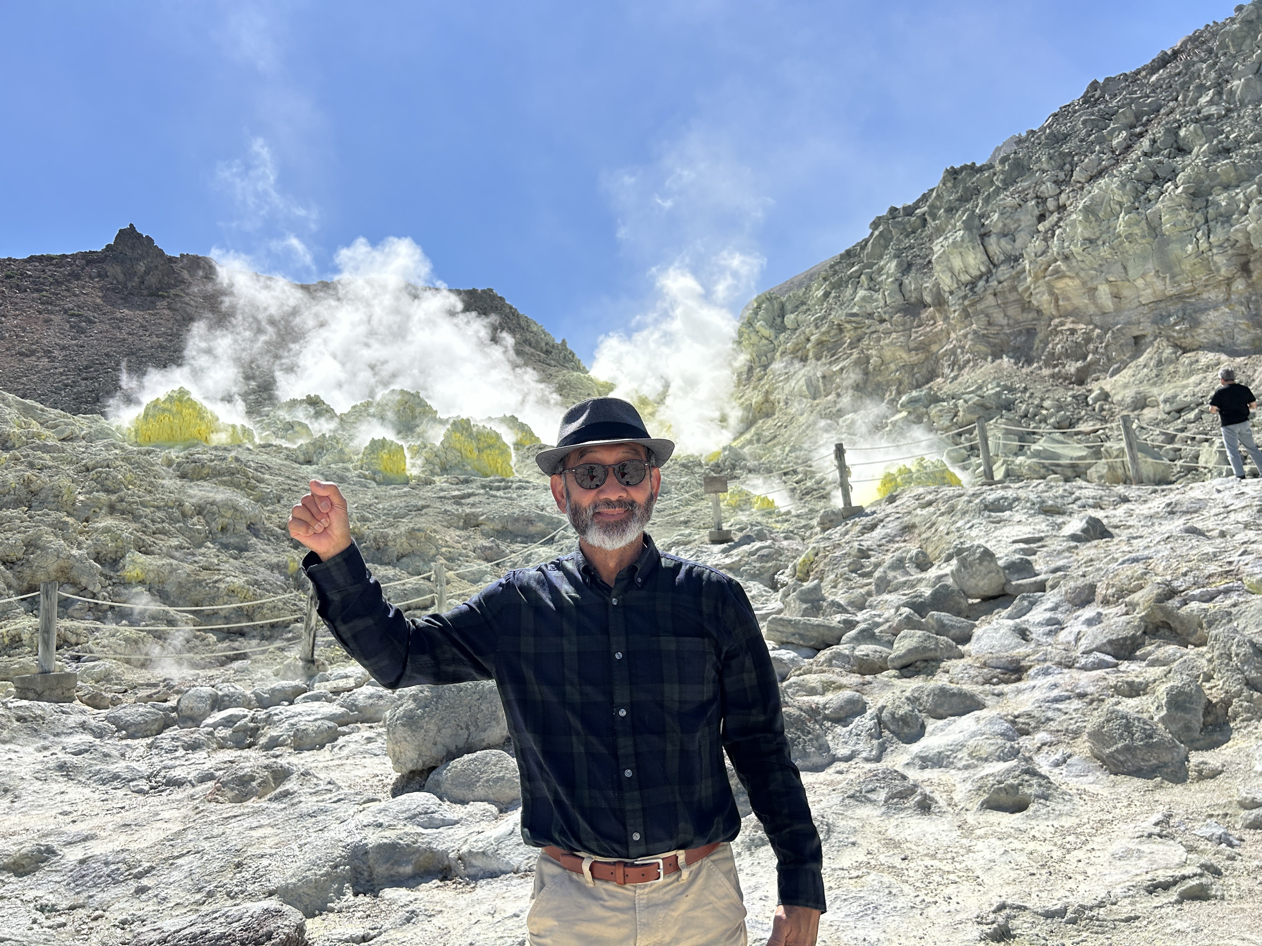 A man stands in front of steaming volcanic vents at Mt. Iozan, Hokkaido, raising a fist in the air and smiling. The rocks behind him are bright yellow from the sulphur.