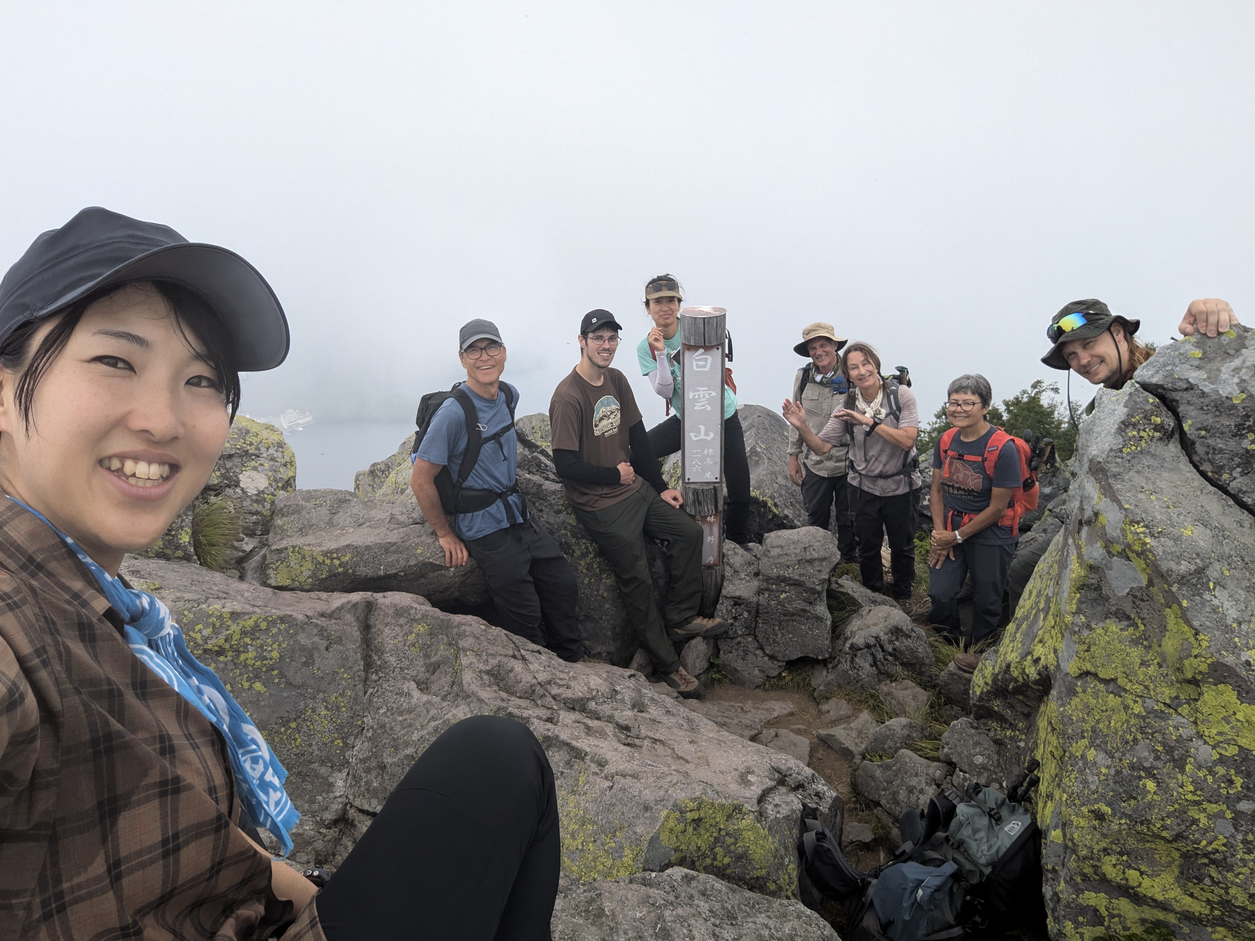 A girl is taking a selfie with a group of hikers at the summit of a mountain. They are all smiling. It is very rocky with large boulders and some people are sitting on them and poking their heads out from behind them. A pole with Japanese text reads "Mt. Hakuunzan, 1186m".