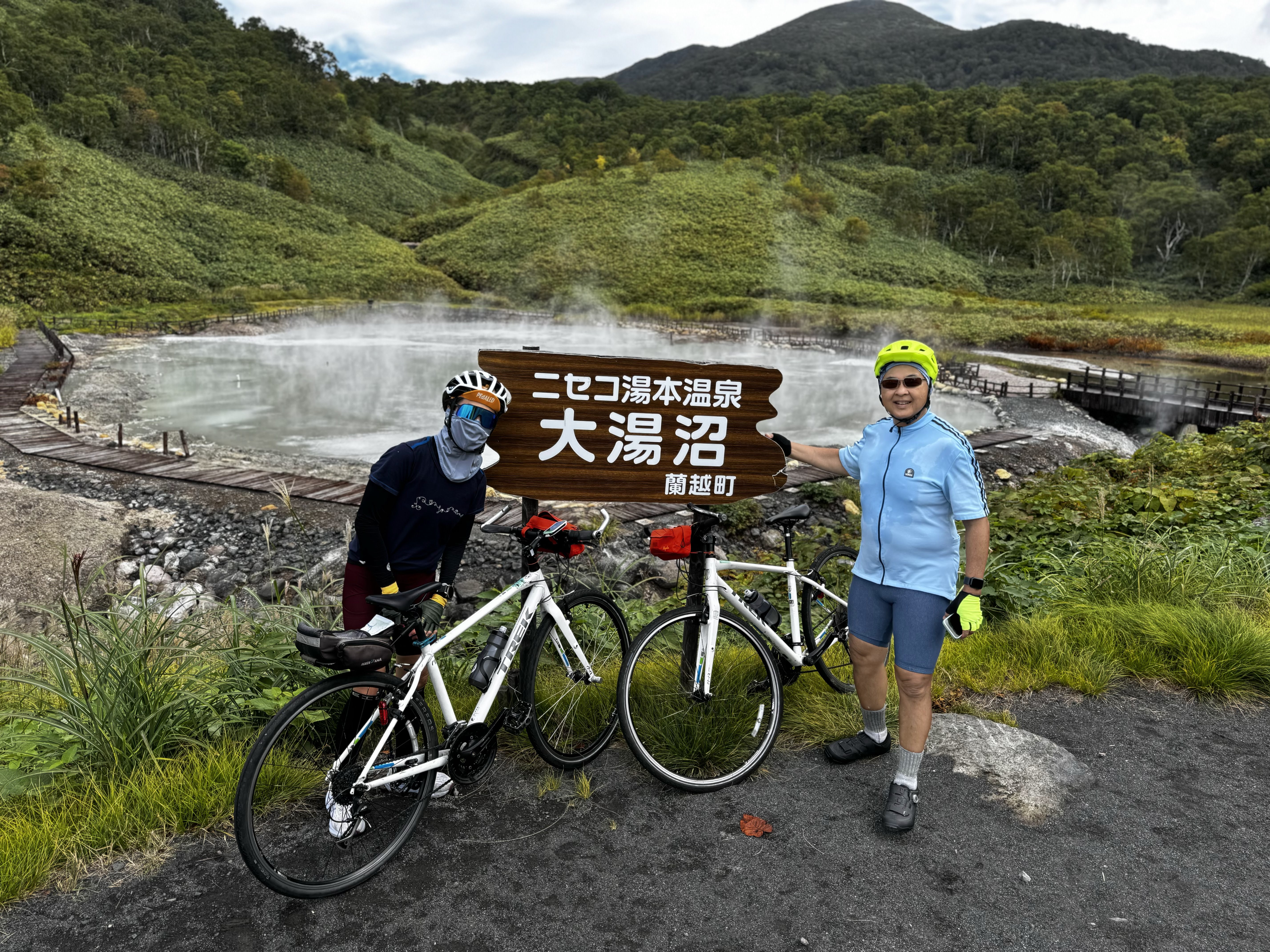 Two cyclists smile with their bikes in front of a sign that reads "Niseko Yumoto Onsen - Oyunuma" in Japanese. Behind them is a hot spring pond, steam rising from its surface.