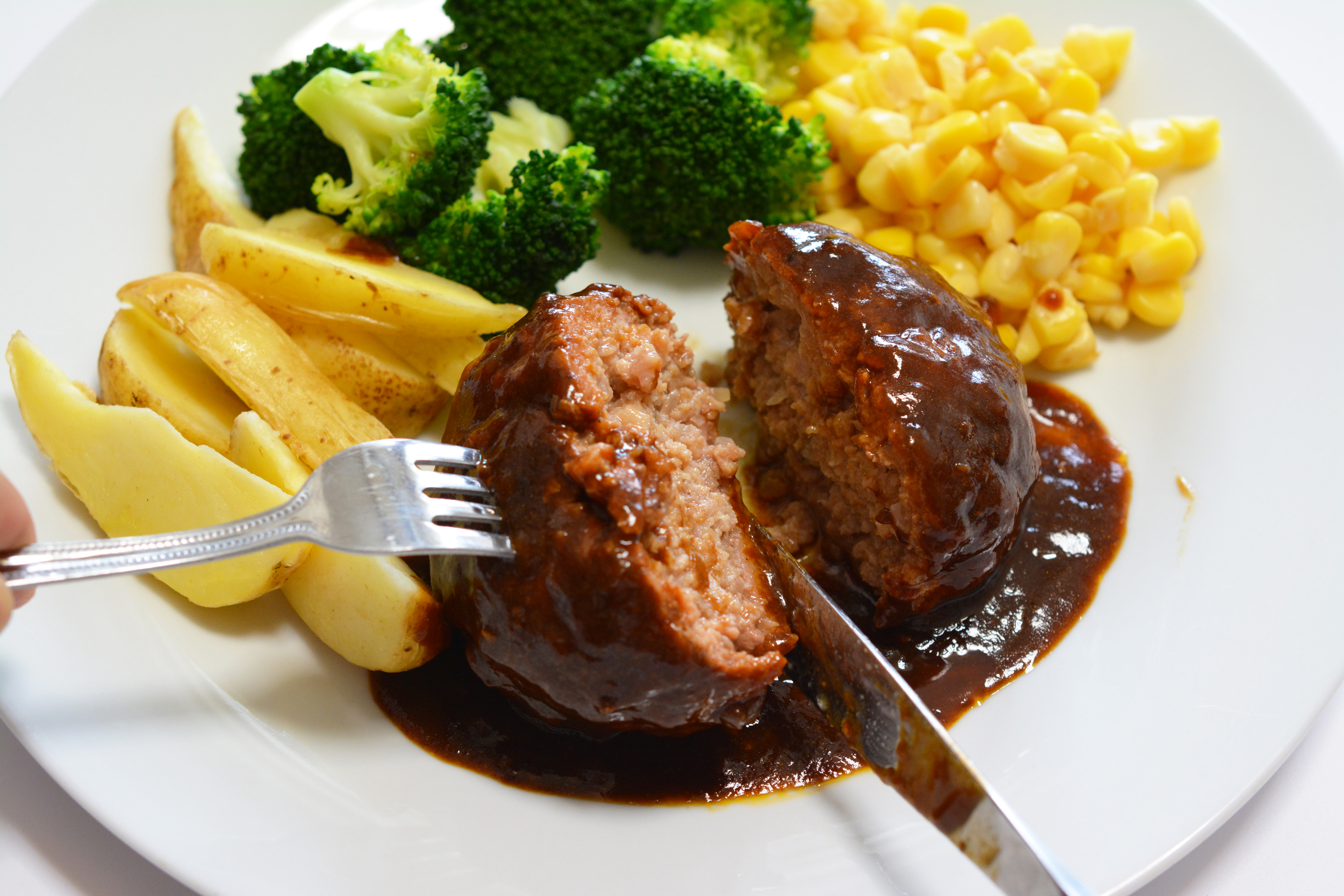 A hamburg steak on a plate with potatoes, broccoli and sweetcorn. A knife and fork are slicing the Hamburg steak in half. The steak is coated with a demi-glacé sauce.