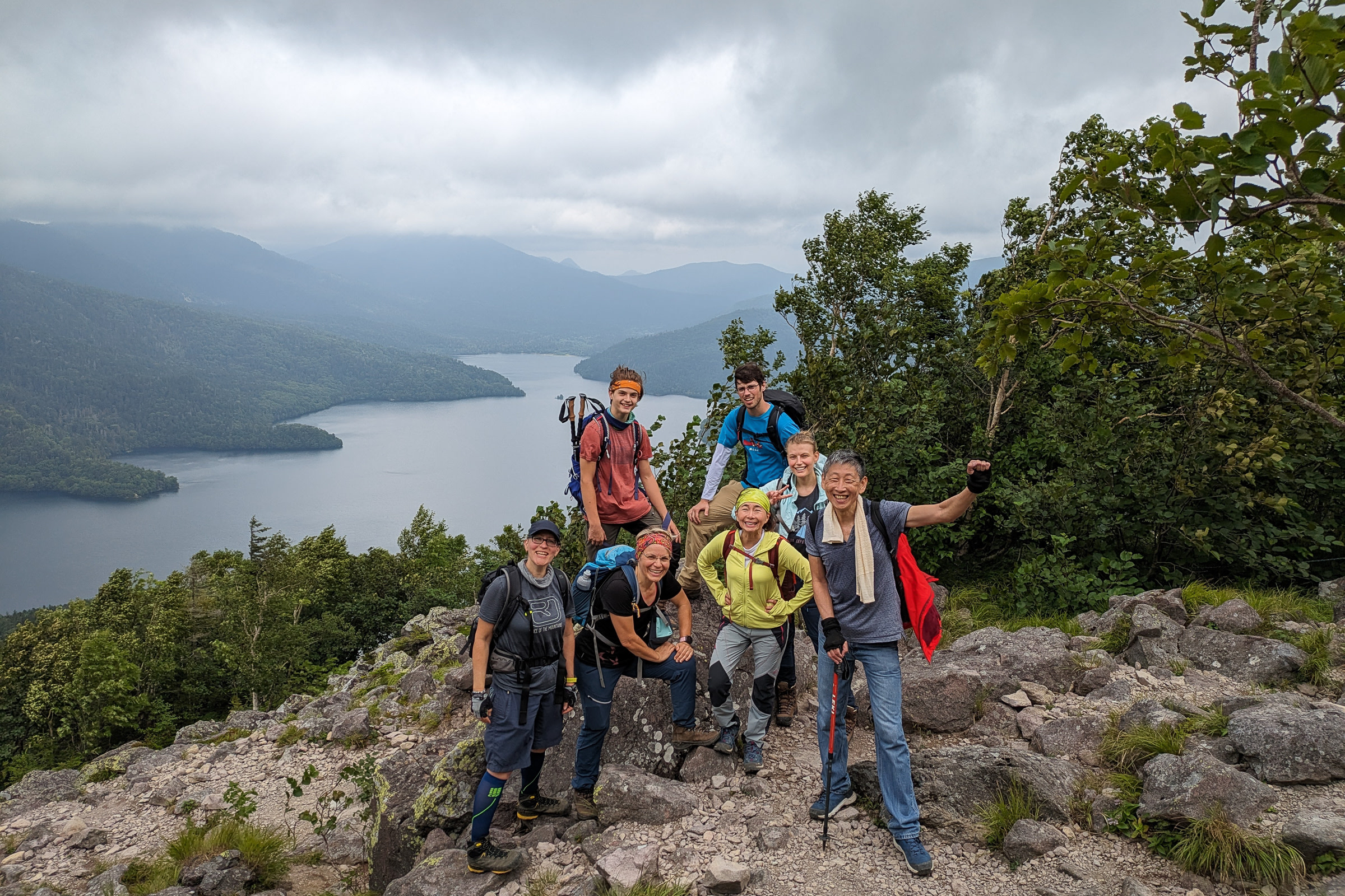 A group of seven hikers beam atop Mt. Hakuunzan, Lake Shikaribetsu in the background. It is a cloudy day.