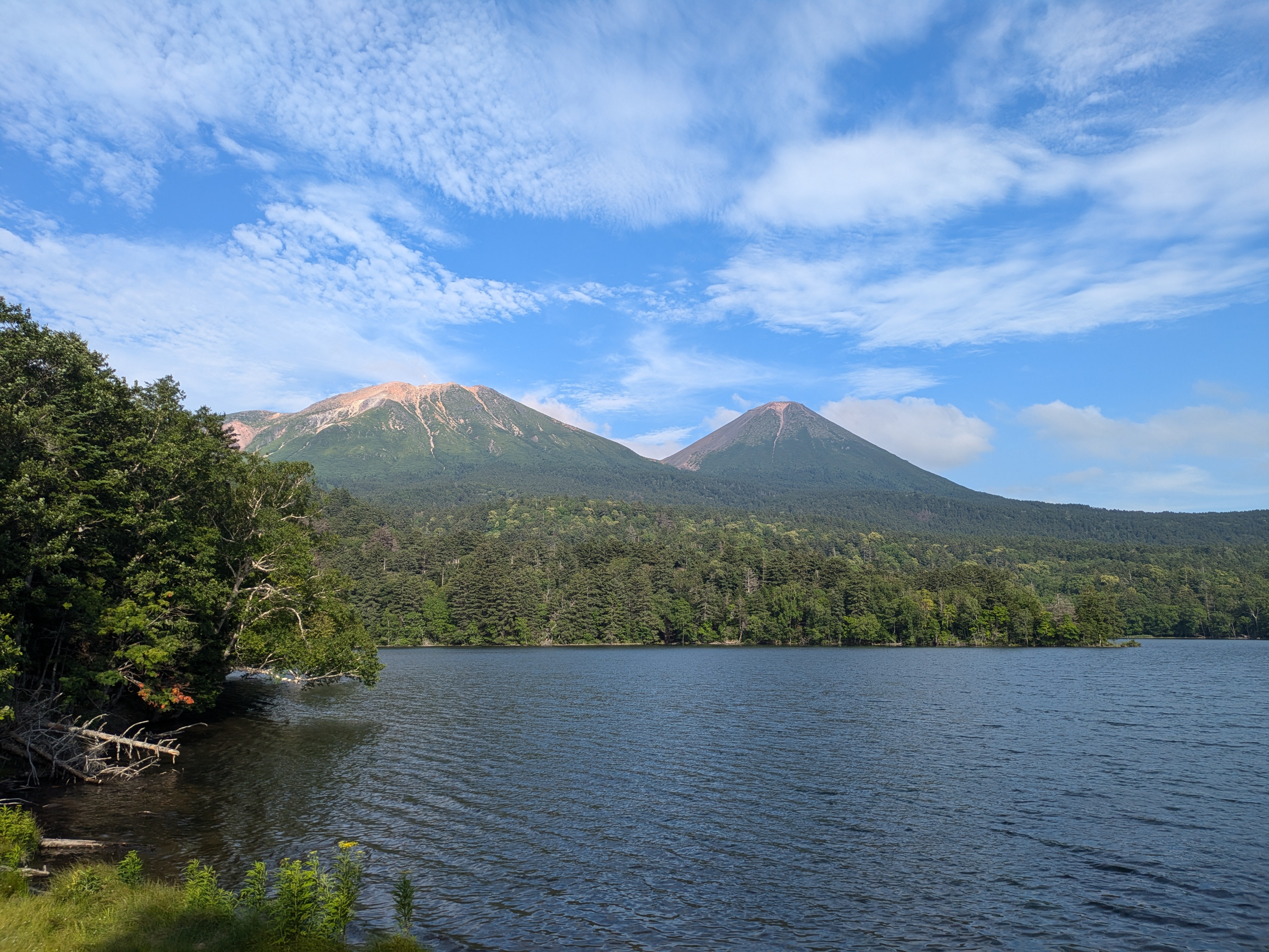 The view of Mt. Meakan and Mt. Oakan from the shoreline of Lake Akan. It is a beautiful day with very clear weather. The lake is surrounded by green trees.