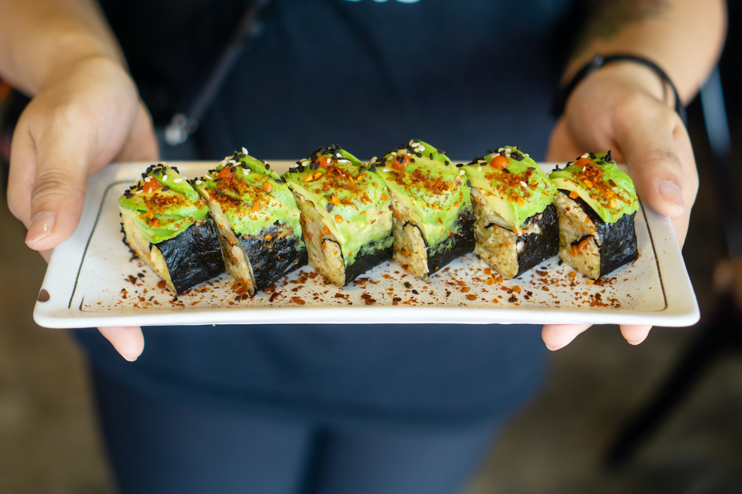 A pair of hands hold a platter of avocado sushi.