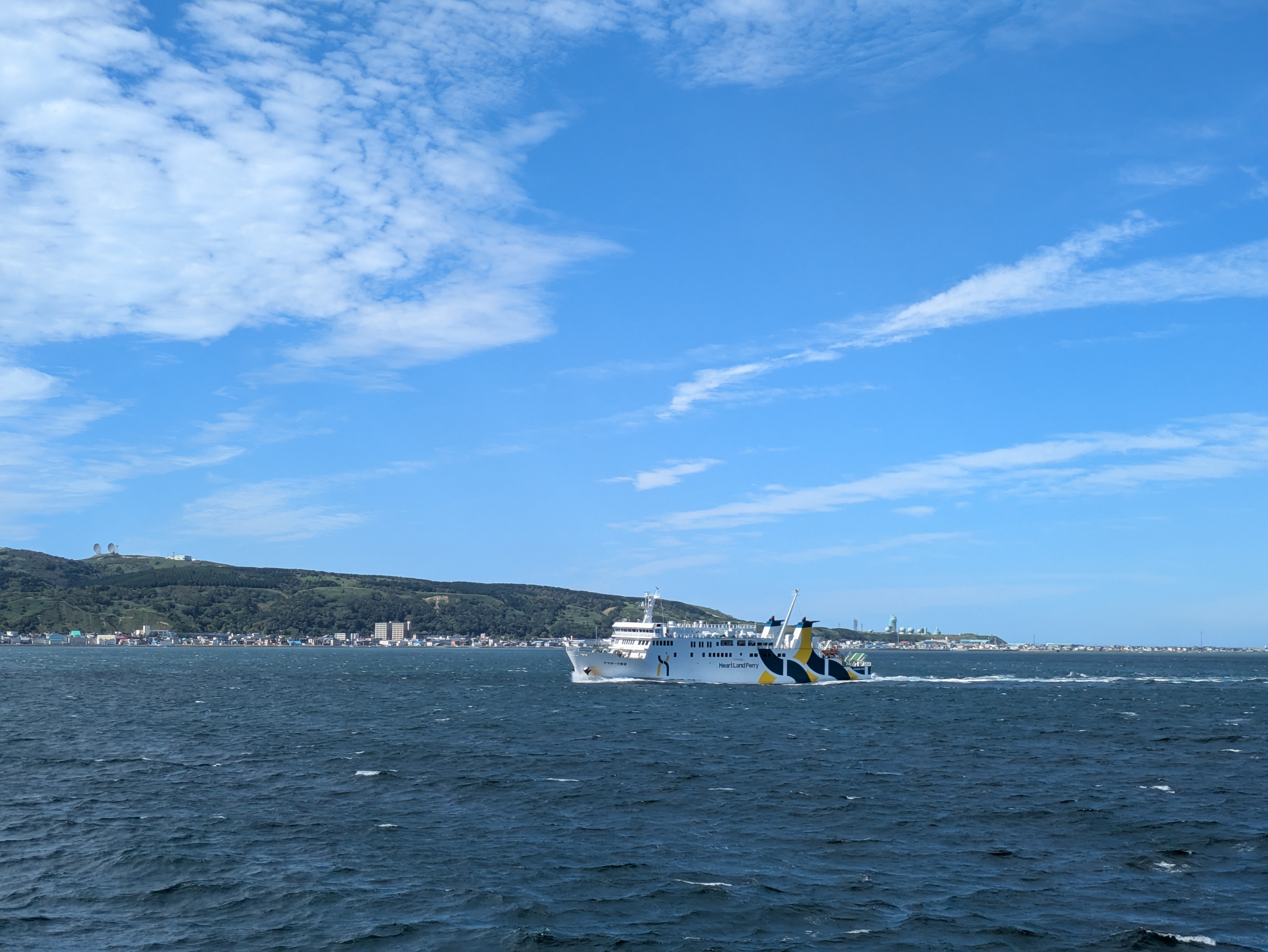 A ferry crosses the ocean in front of an island in the distance. It is a beautiful sunny day. The boat is labelled with the words "Heart Land Ferry".