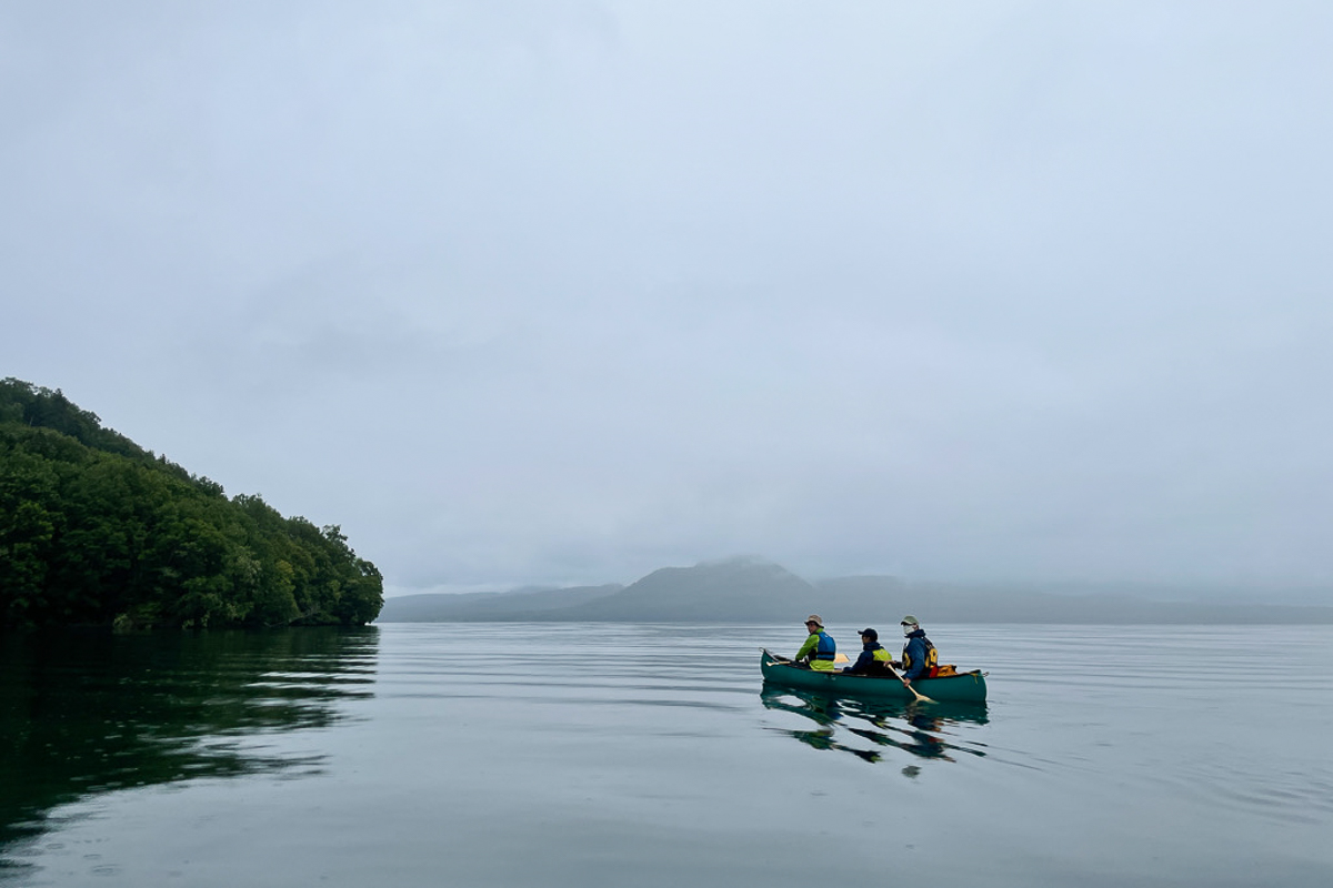 Three people float in a canoe on Lake Kussharo on a cloudy and still day.