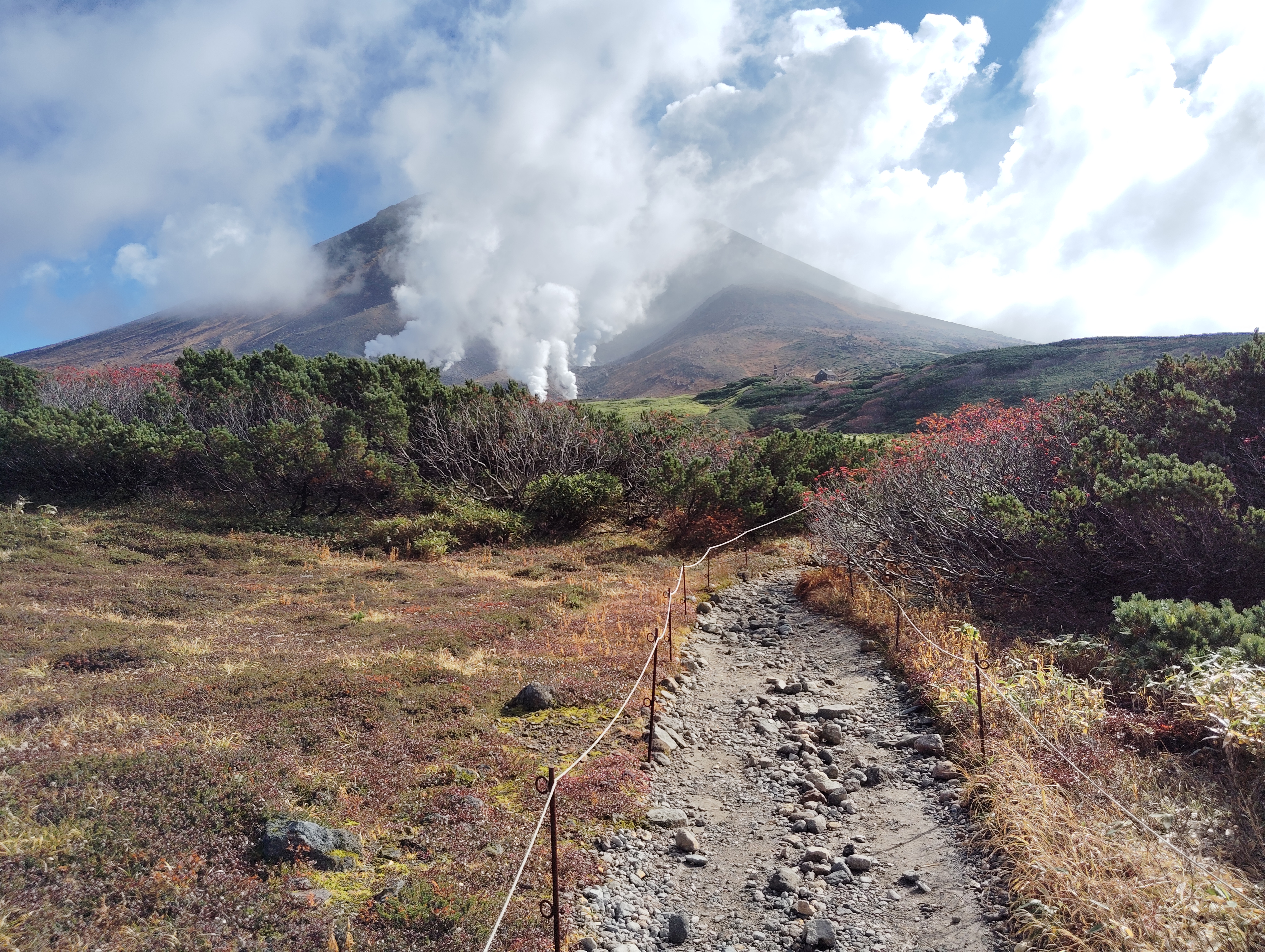 A view of Mt. Asahidake in autumn. The grasses and bushes around the rocky trail are orange, red, brown and yellow with autumn. In front of the mountain, volcanic fumaroles send up powerful columns of steam into the air.