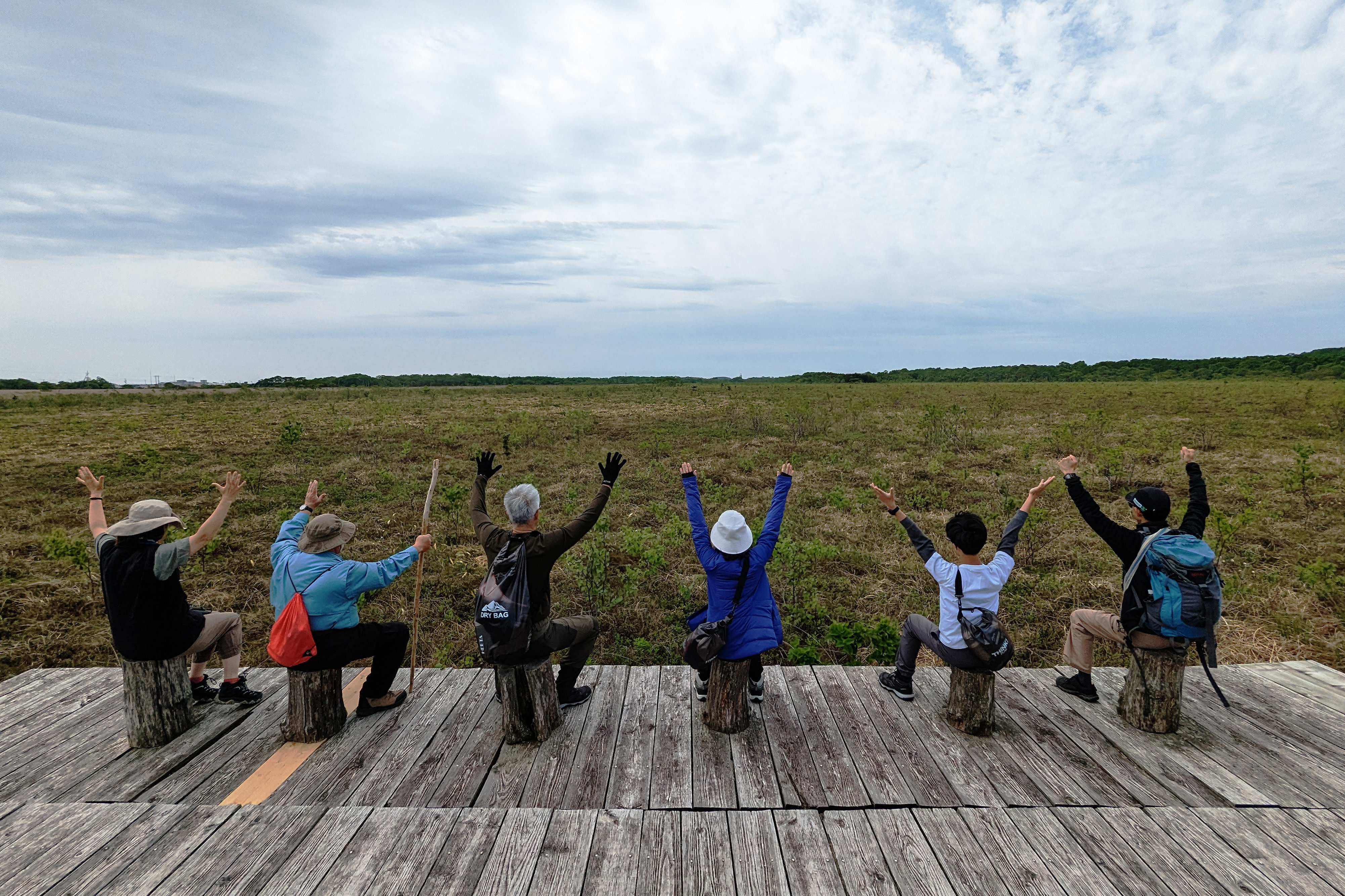A group of walkers pose for a photo with everyone sat on wooden stools. The group faces away from the camera and towards a wetland