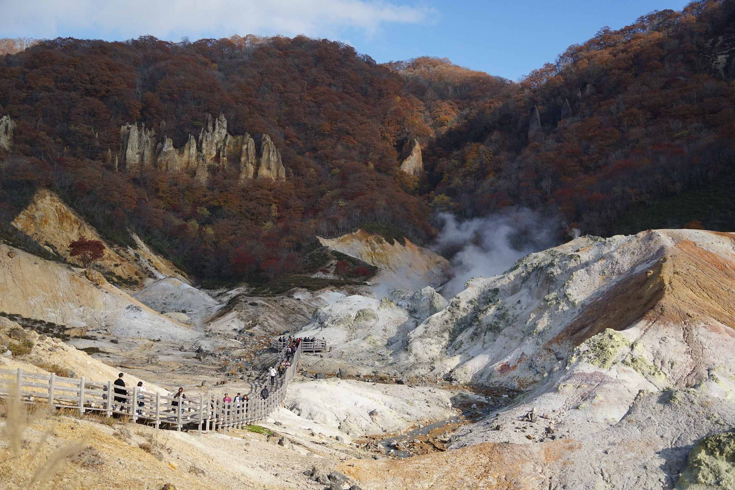 A view into Noboribetsu Hell Valley. A wooden walkway takes hikers down into the valley, steam from volcanic vents rising all around. The landscape looks sandy and rocky.