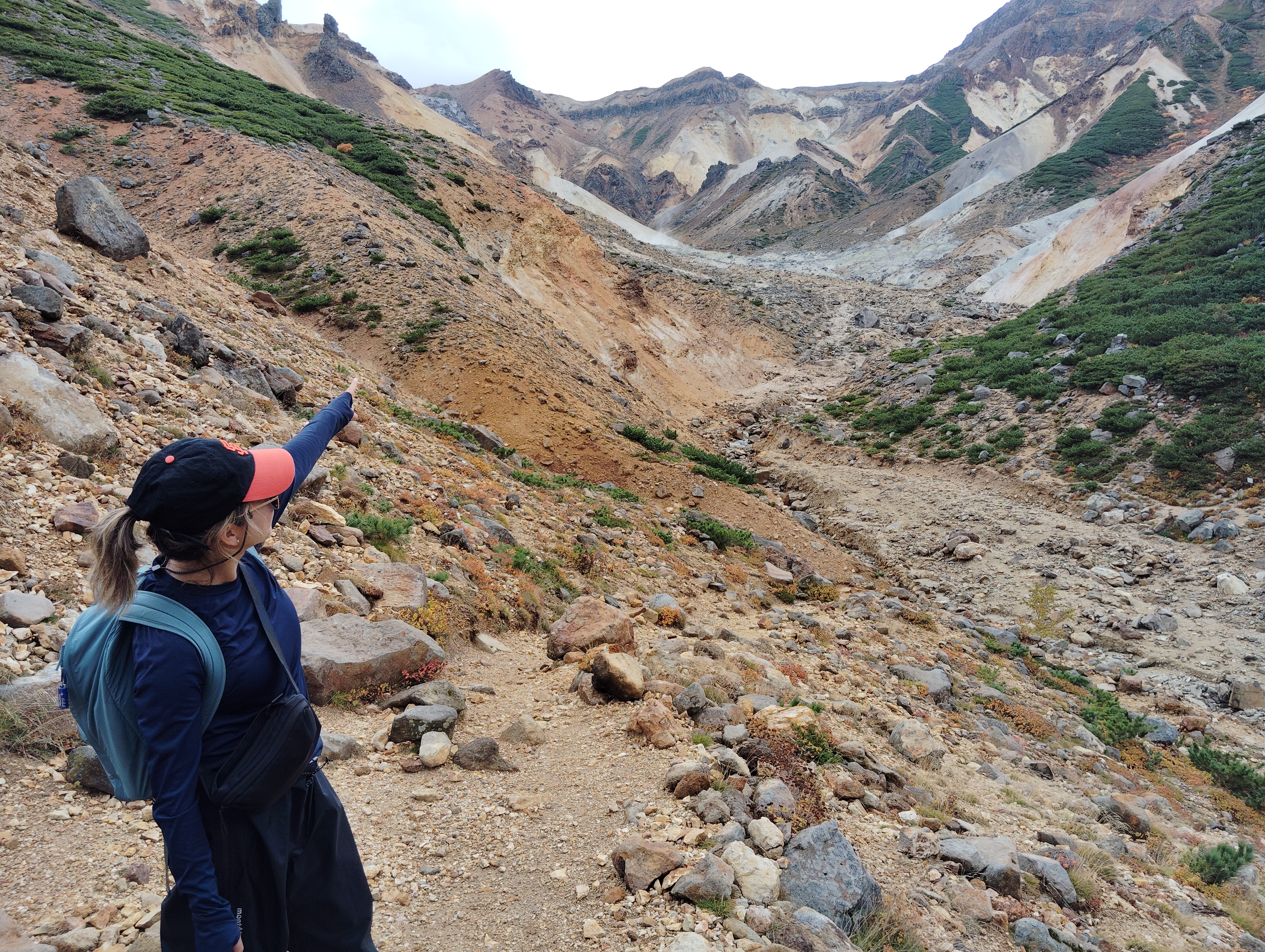 A woman stands in a rocky mountain valley on Mt. Tokachi, Hokkaido. She is pointing to the rocky and sandy slopes of a mountain in the middle distance.