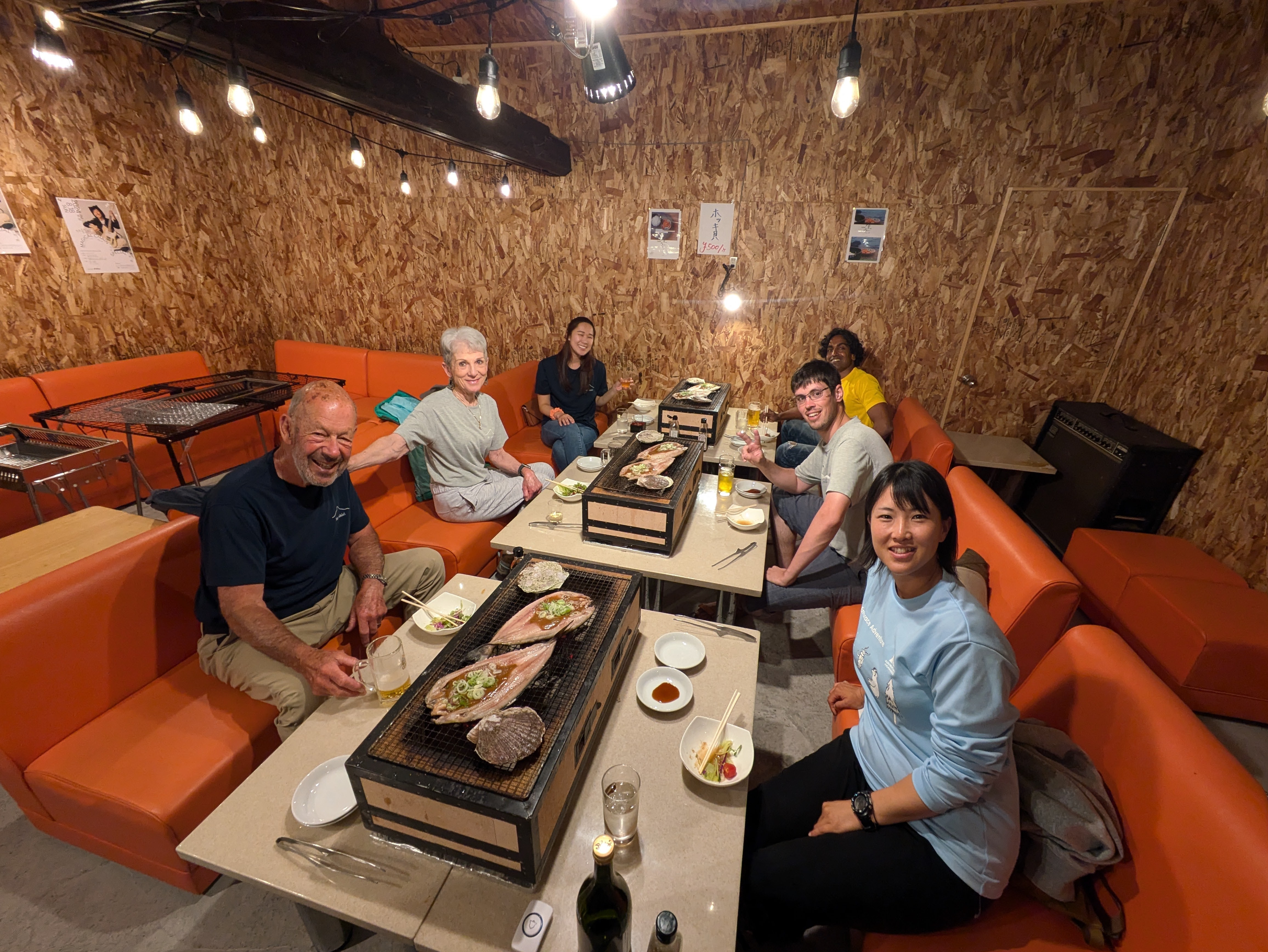 A group of diners sit spread across three tables, smiling at the camera. There are grills perched on the tables and butterflied fish are laid out on the grill.