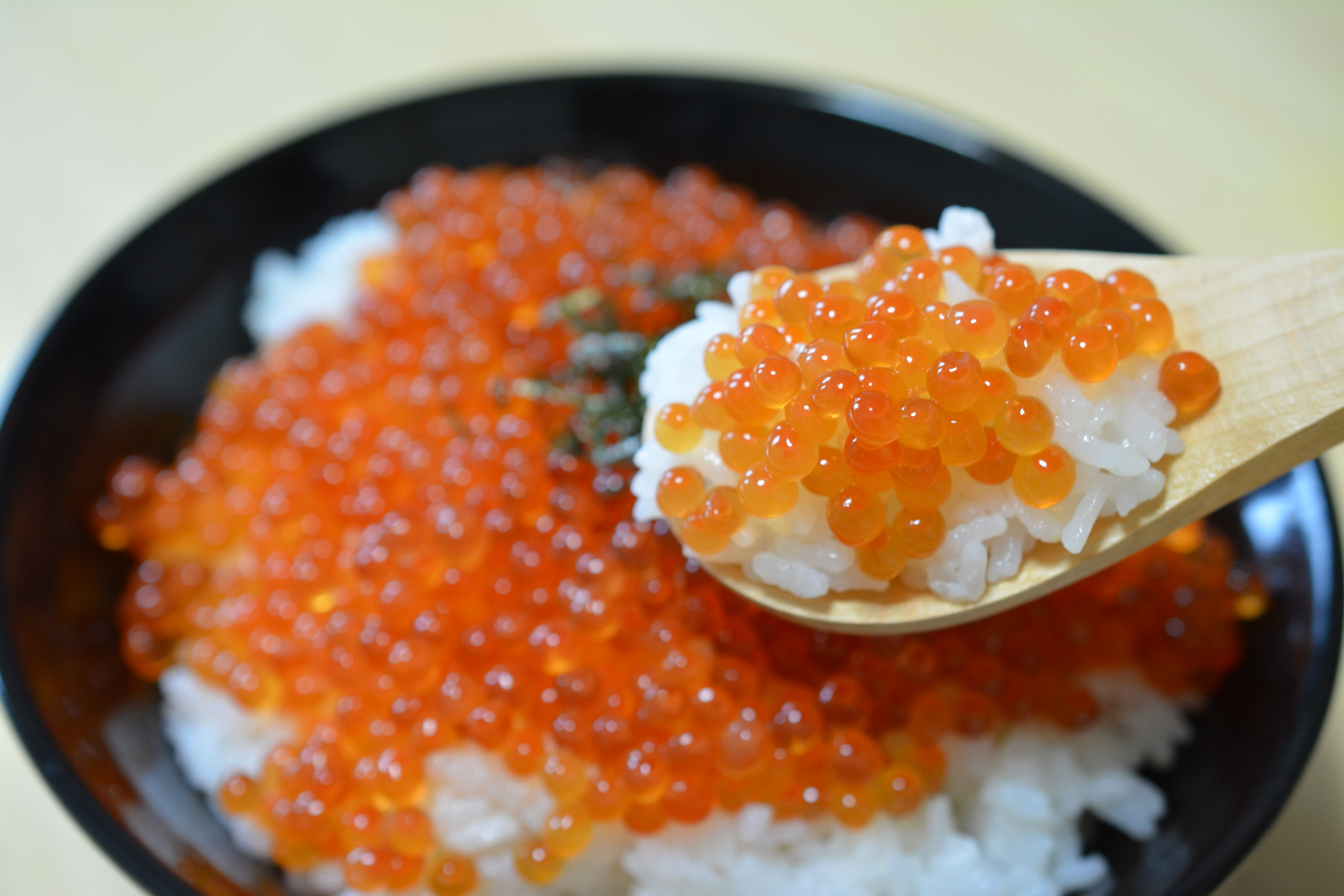 A rice bowl with a topping of salmon roe. In the foreground, a wooden spoon takes a scoop of the rice with some salmon roe on top.