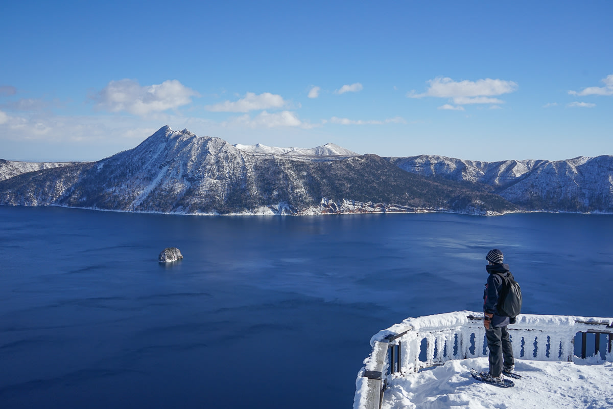 A person wearing snowshoes looks out over the deep blue waters of Lake Mashu. It is midwinter, and the lookout point is frozen over.
