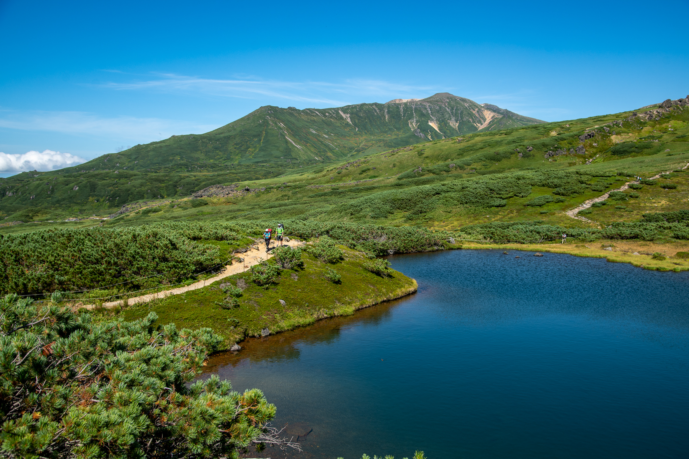 A still pond is surrounded by greenery. Two hiker walk on a trail along the ponds edge.