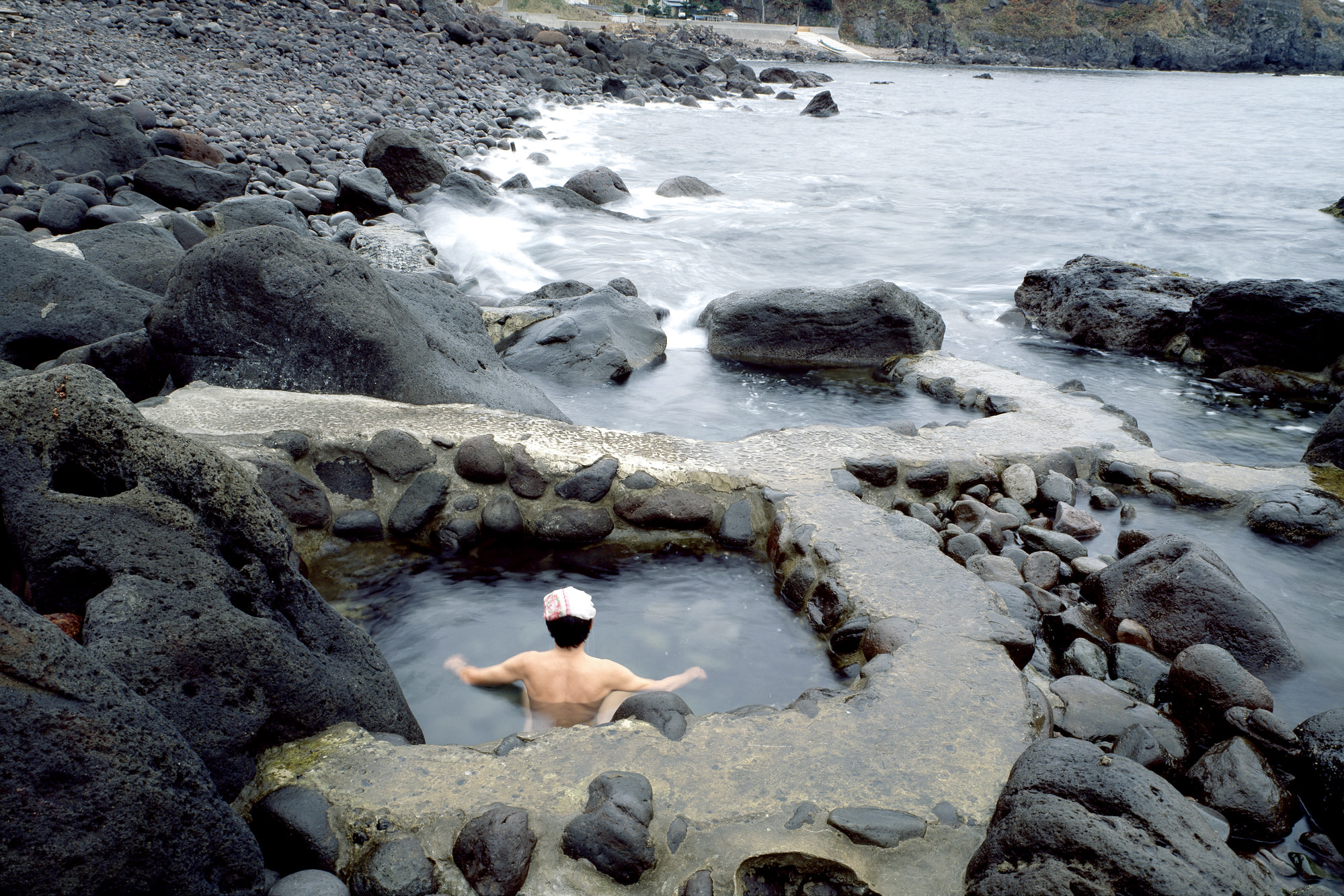 A man soaks in a small onsen hot-spring pool next to the sea. Waves can be seen crashing on the shore in the distance.