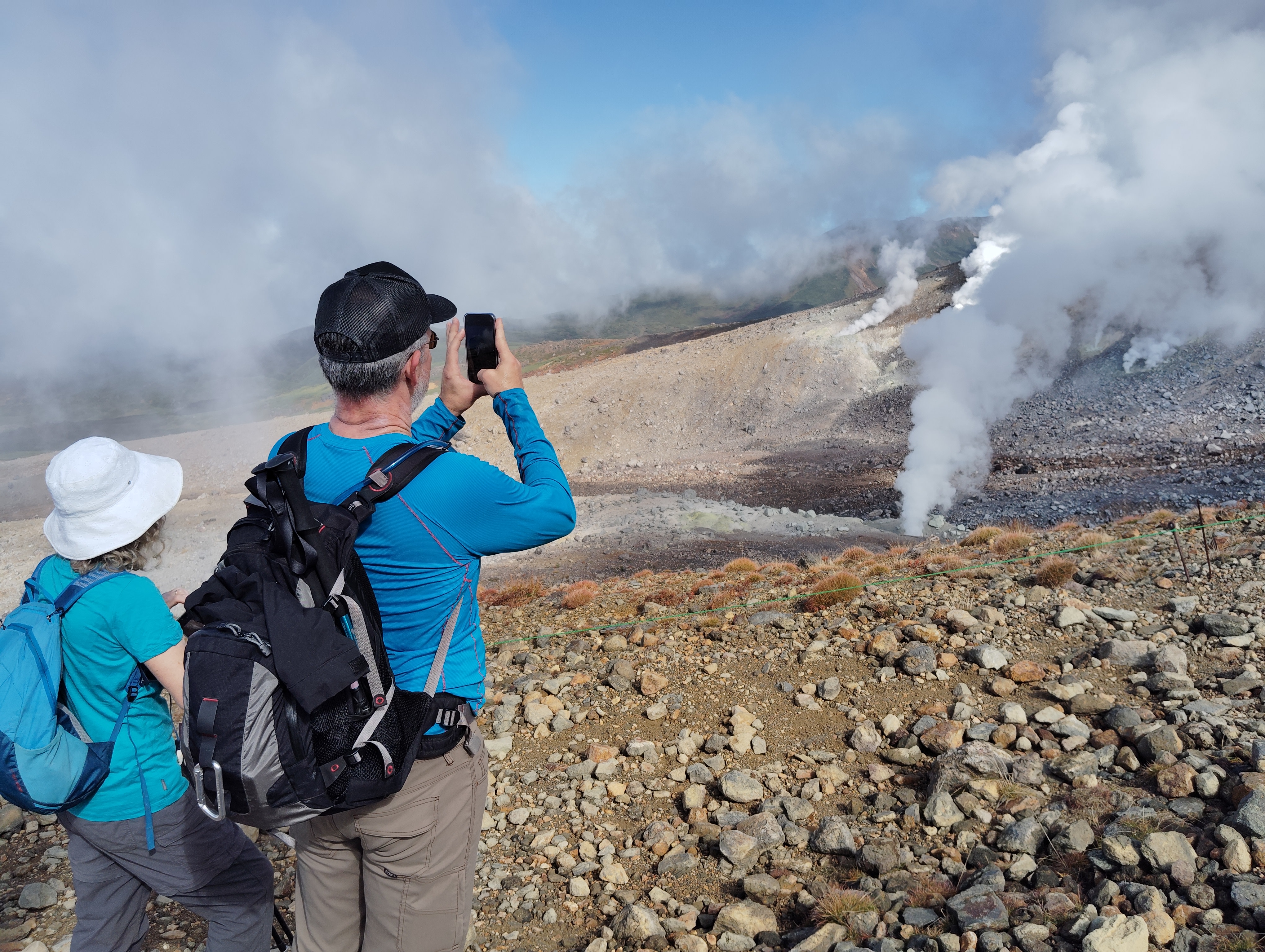 Two hikers, a man and a woman, observe volcanic fumaroles on Mt. Asahidake, Hokkaido. Steam rises powerfully from the vents in large columns. The man is taking a photo of the sight on his smartphone.