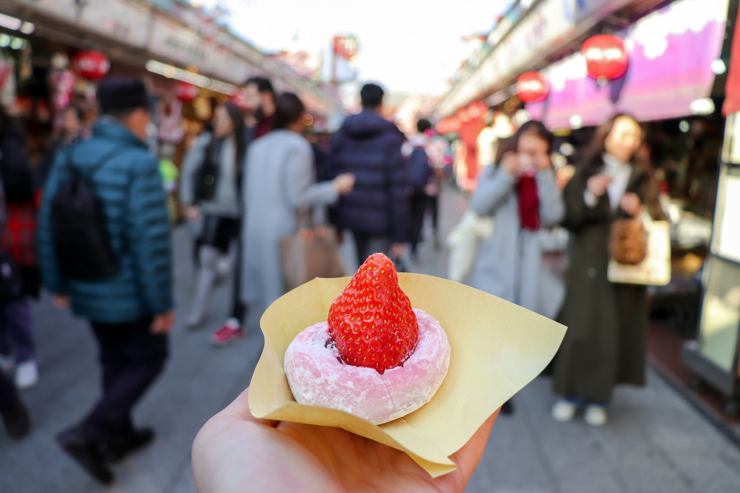 A hand holds a strawberry Saifuku (rice cake filled with sweet bean paste) in the middle of a crowded shopping street.