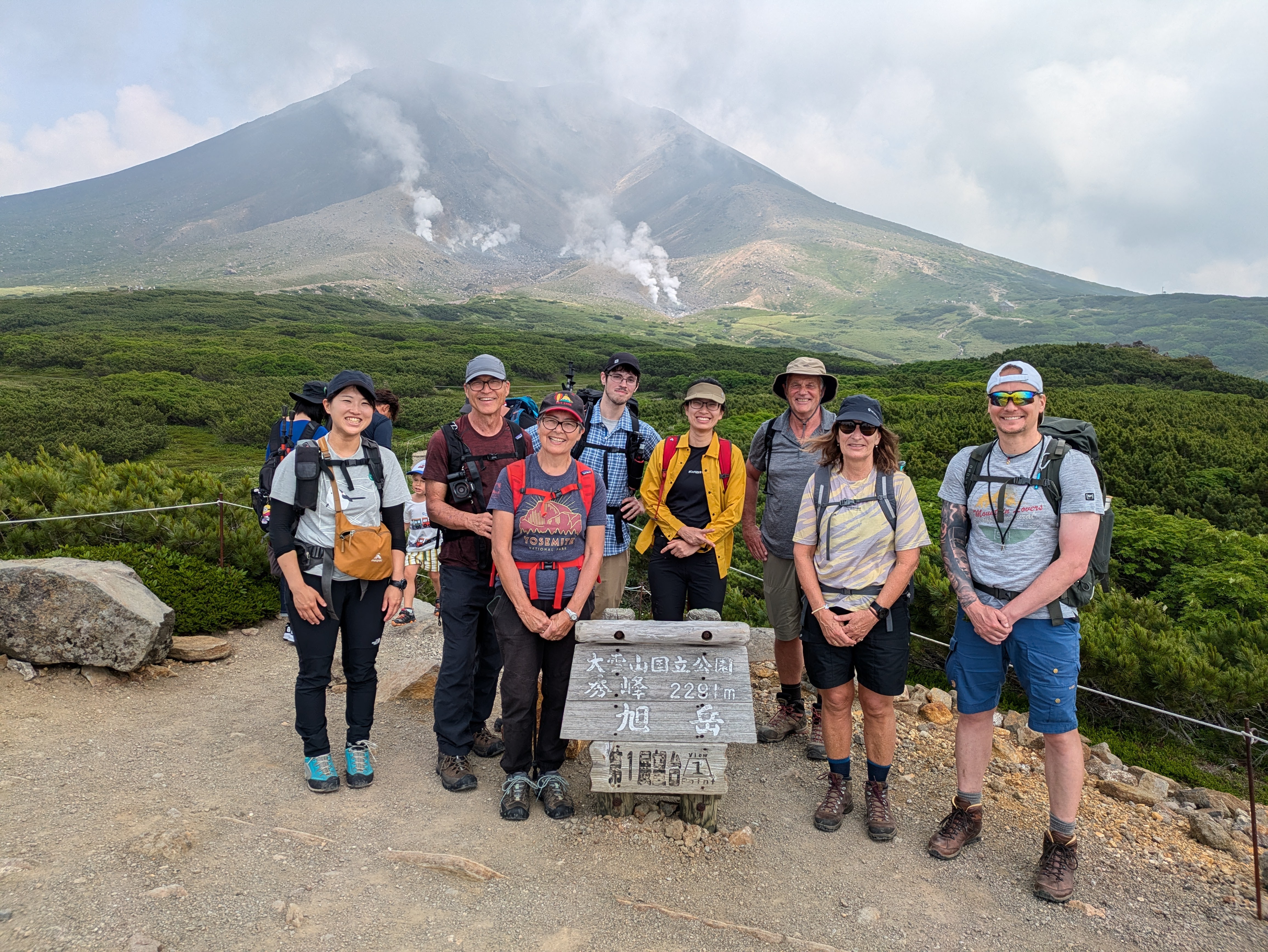 A group of hikers smile at the camera with Mt. Asahidake, the tallest mountain in Hokkaido, behind them. Steam is visibly rising from volcanic vents on the mountain.