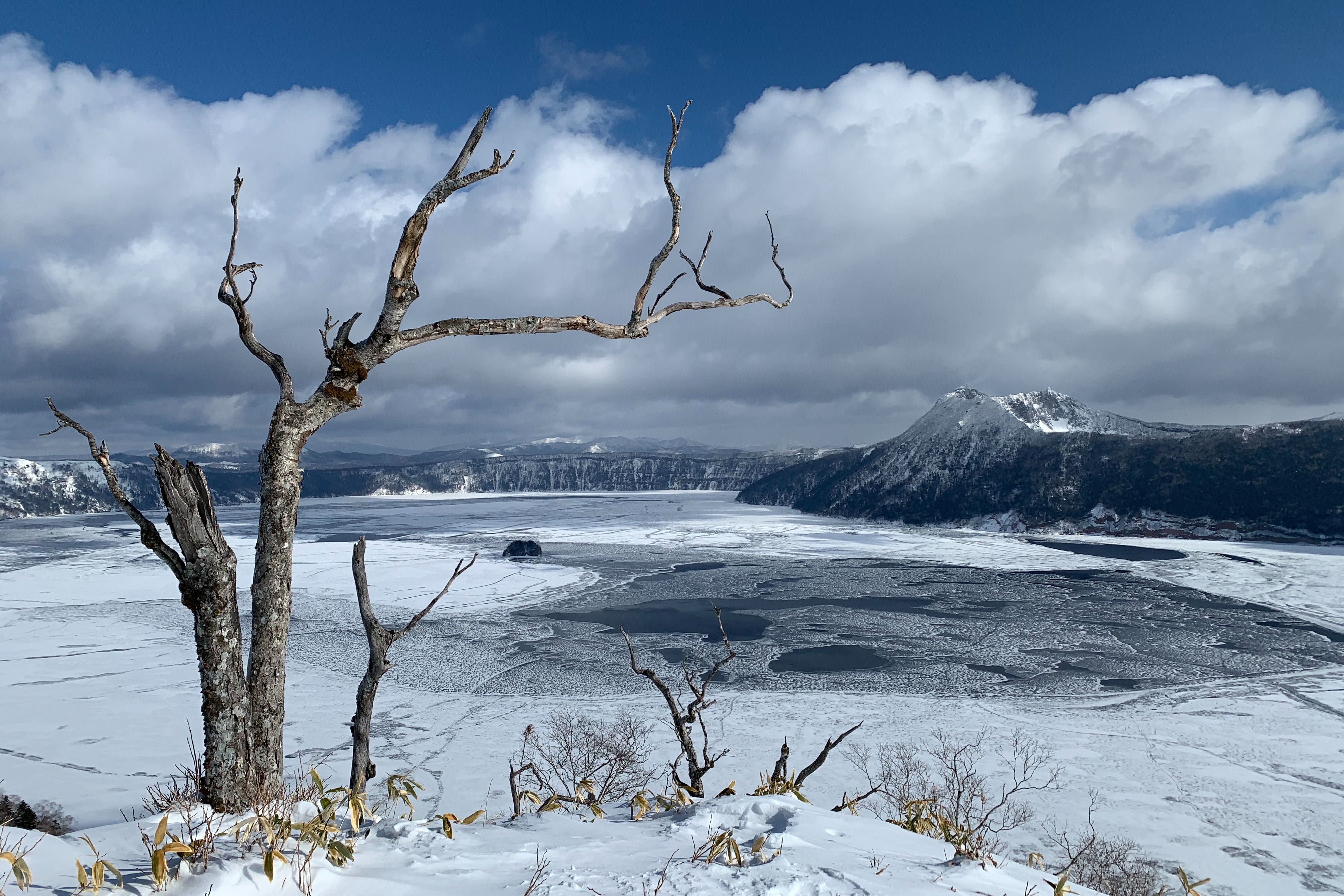 A view of Lake Mashu in winter from a view point. The surface of the lake is covered in big plates of ice. It is a beautiful day with blue sky above.