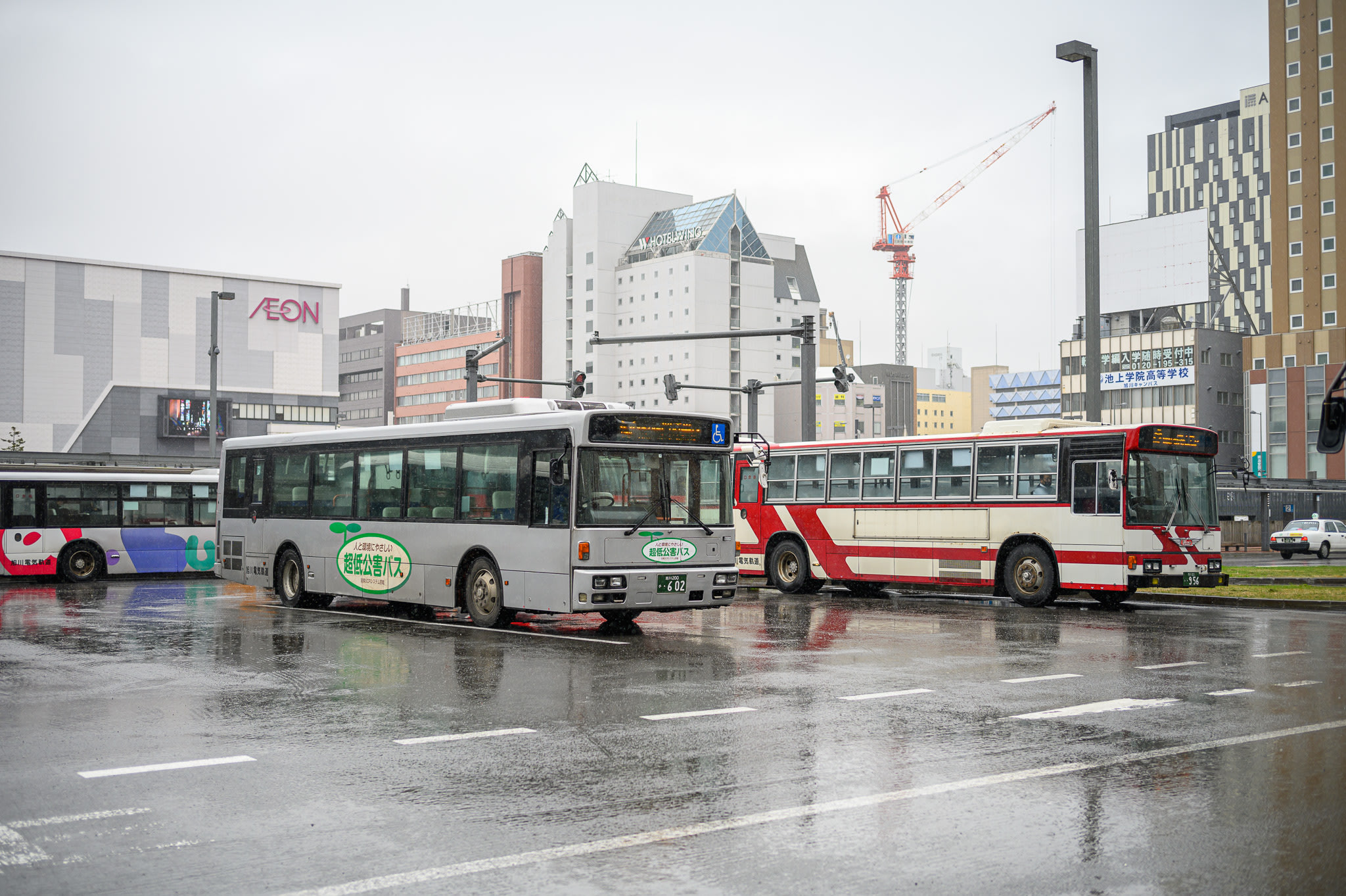 Two local buses parked at the bus stops in front of Asahikawa Station on a rainy day.
