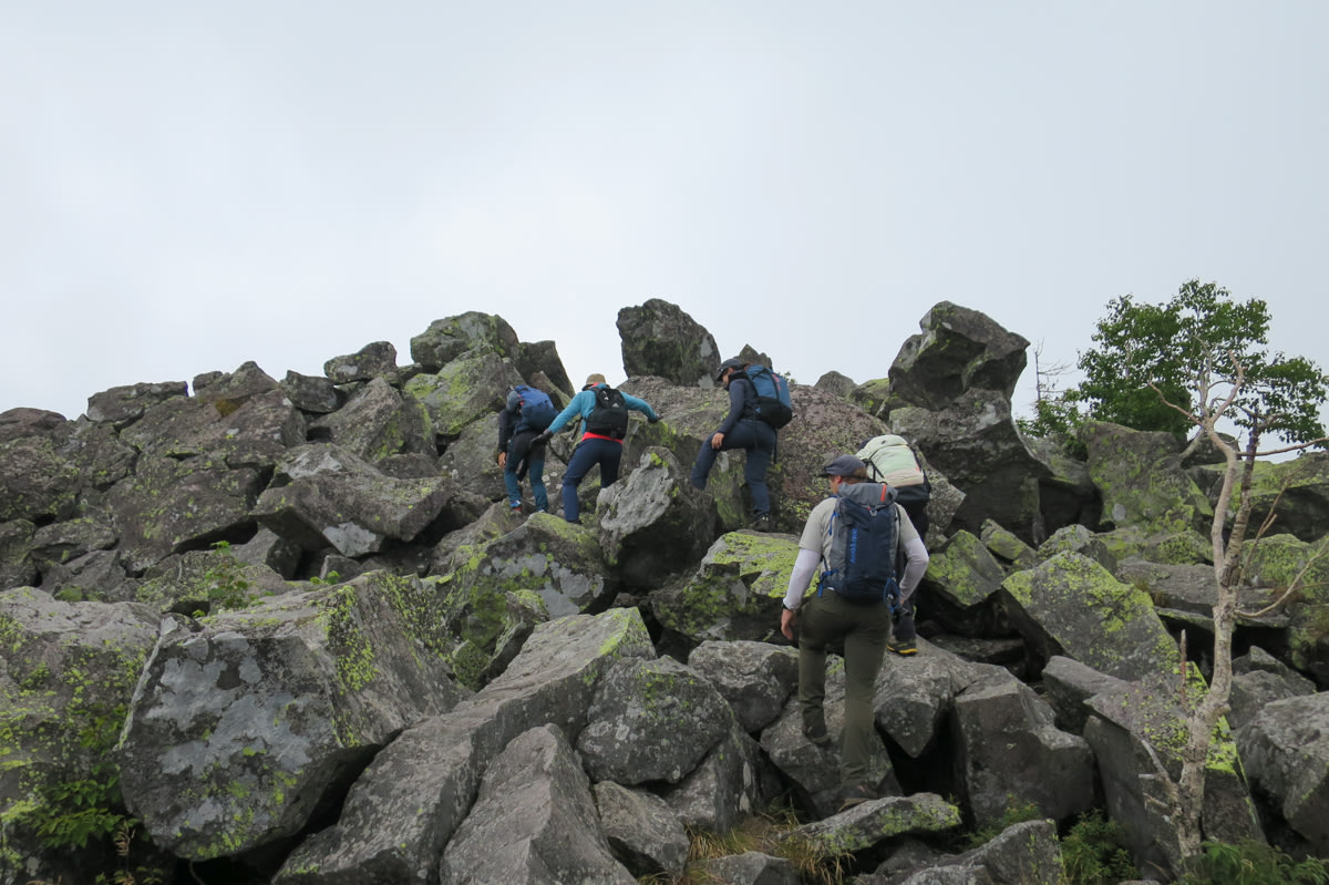 A group of hikers pick their way over many large fallen boulders, piled up to the horizon.