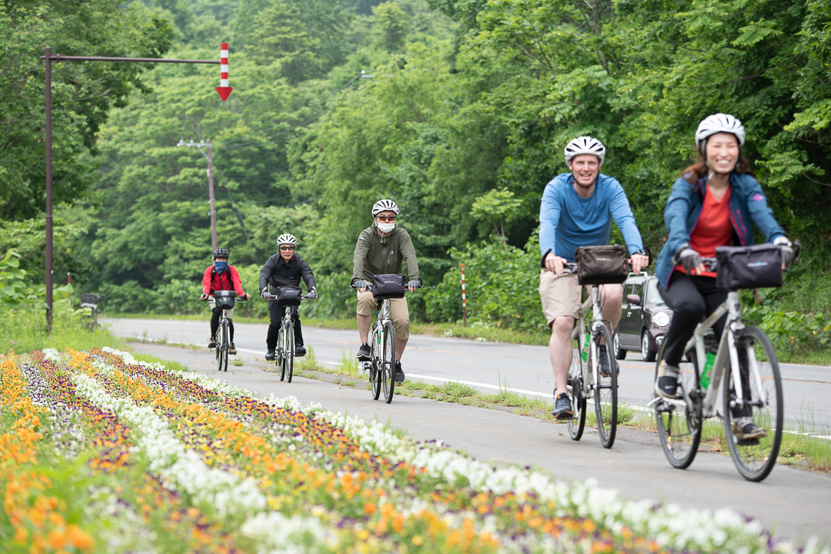 Five cyclists ride by a field of bright flowers.