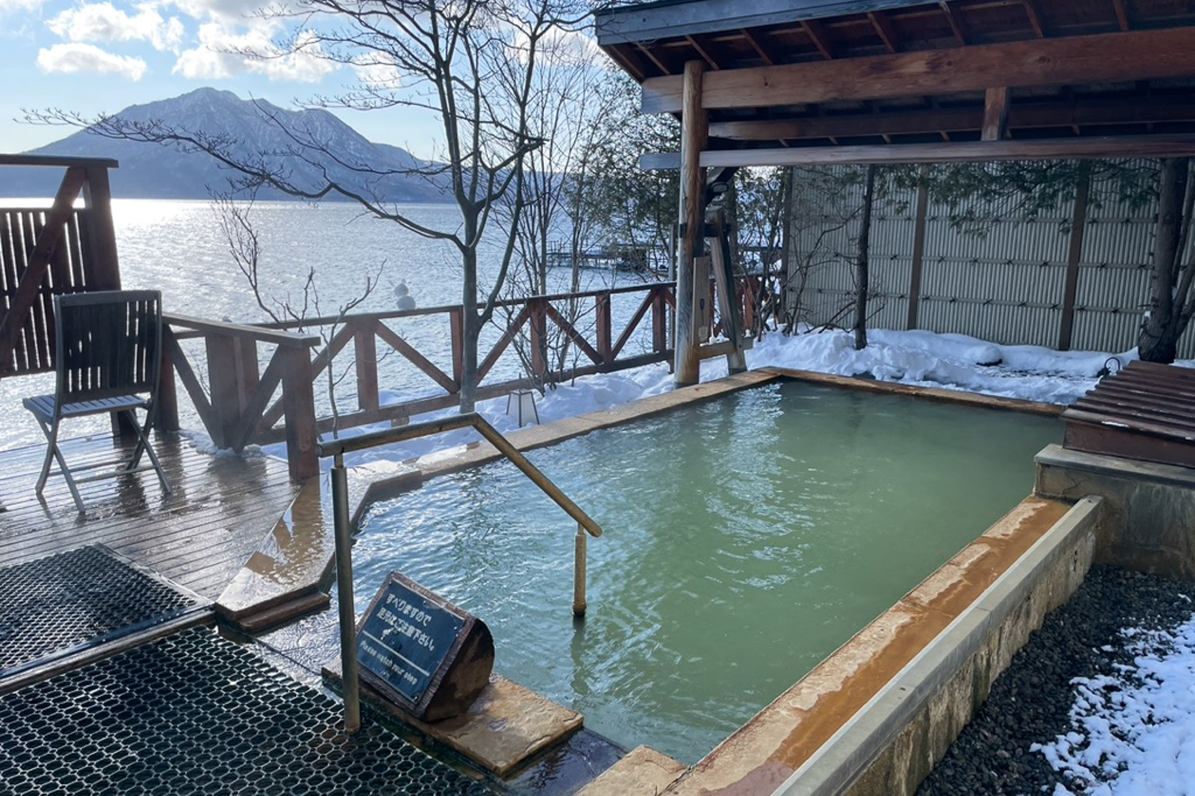 A routenburo Japanese outdoor hot spring pool is surrounded by snow. The pool has a steep handrail leading into it and a gazebo cover at the far end. There is a non-slip rubber mat near the entrance. Behind the pool is a single seat and a wooden fence. Beyond the fence you can see Lake Shikotsu, with the sun reflecting on the lakes surface, and Mt. Fuppushi.