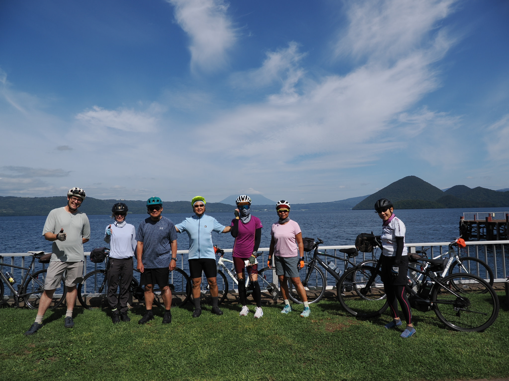 A group of seven cyclists stand in front of a fence at Lake Toya, Hokkaido. They all smile at the camera. It is a very sunny day.