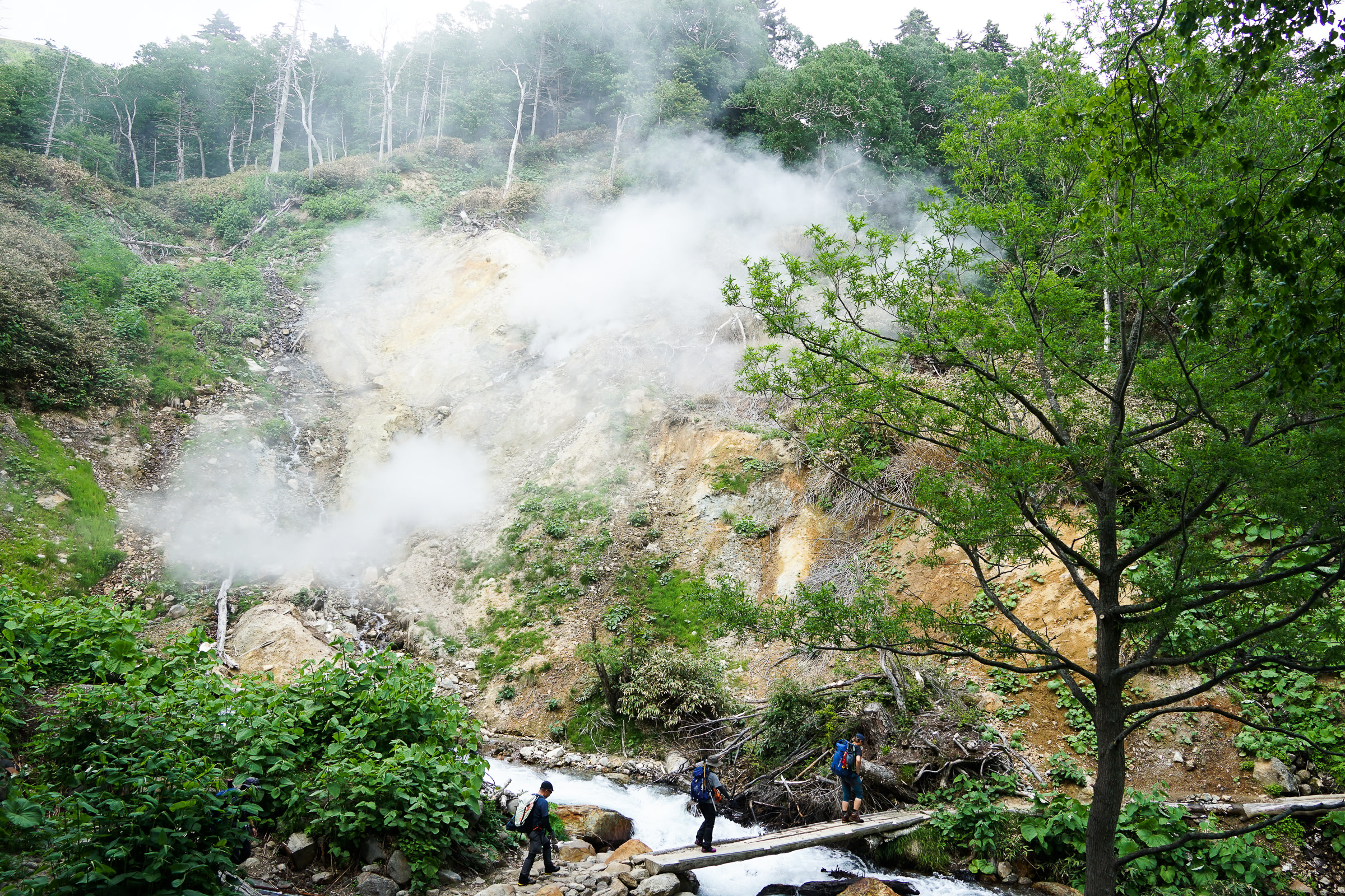 Hikers cross a stream at Kogen Numa while steam billows out of a fumarole.