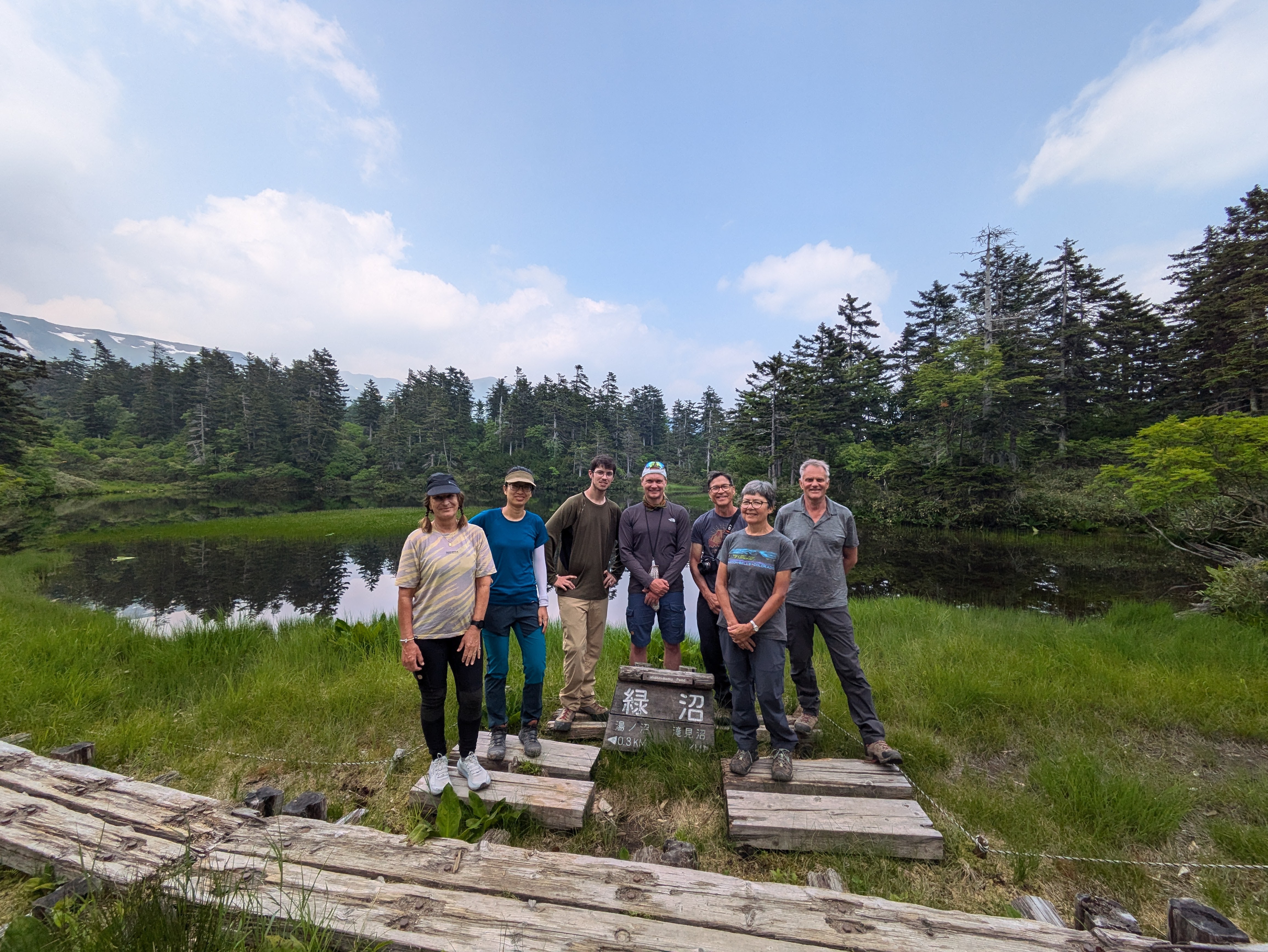 A group of smiling hikers surround a sign reading "Midorinuma" in Japanese. There is an alpine tarn (a pond) behind them. The pond is surrounded by trees. The weather is very clear and still.