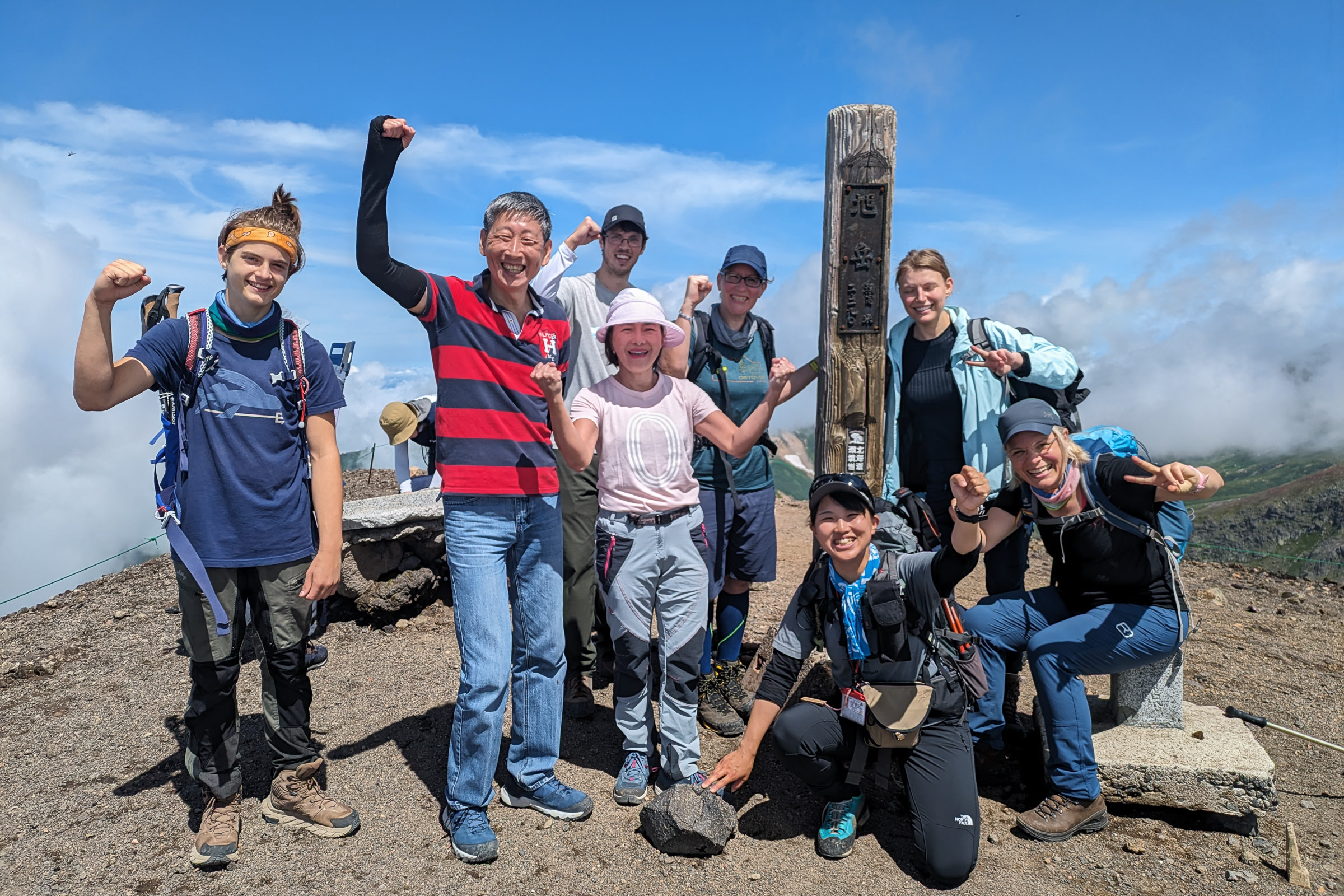 A group of eight hikers smile at the summit of Mt. Asahidake on a sunny day. A pole in Japanese reads "Mt. Asahidake". 