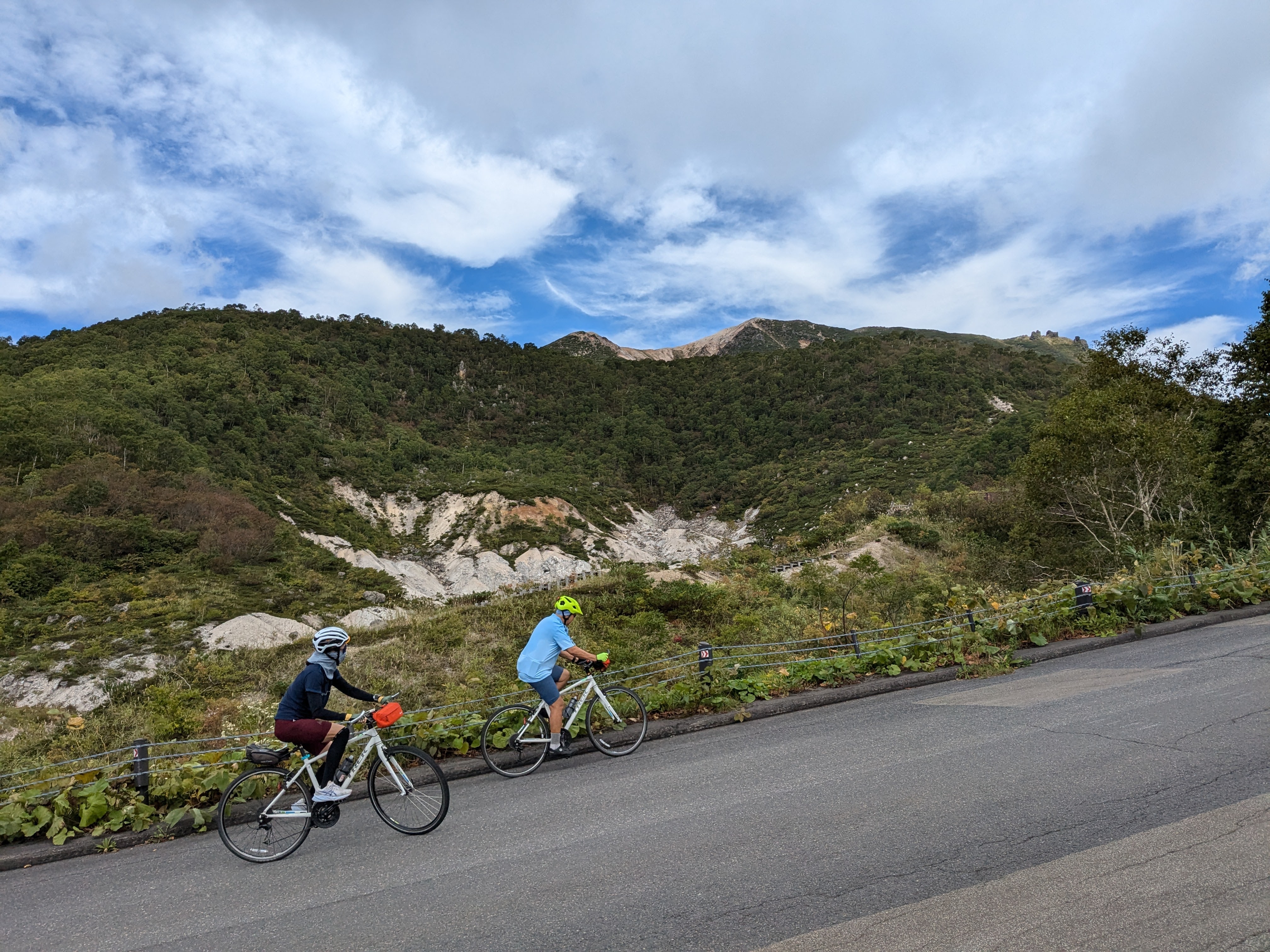 Two cyclists pass by a volcanic-looking crater on the side of the road in a mountain pass. The sky is cloudy, but clearing.