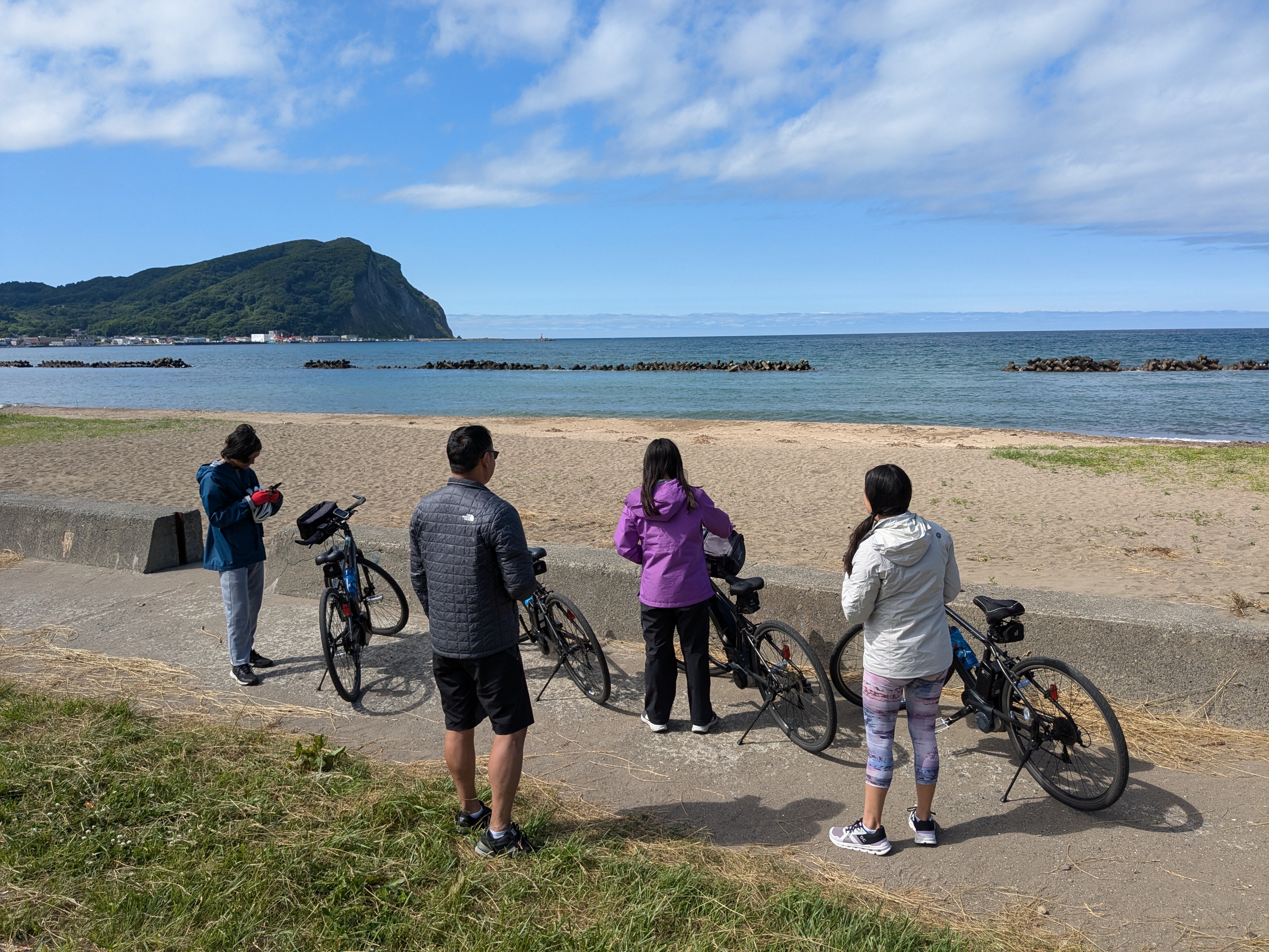 A group of four cyclists looks out at a beach and the sea of Japan from behind a coastal wall. They have their bikes with them, but they are dismounted. It is a very sunny day.