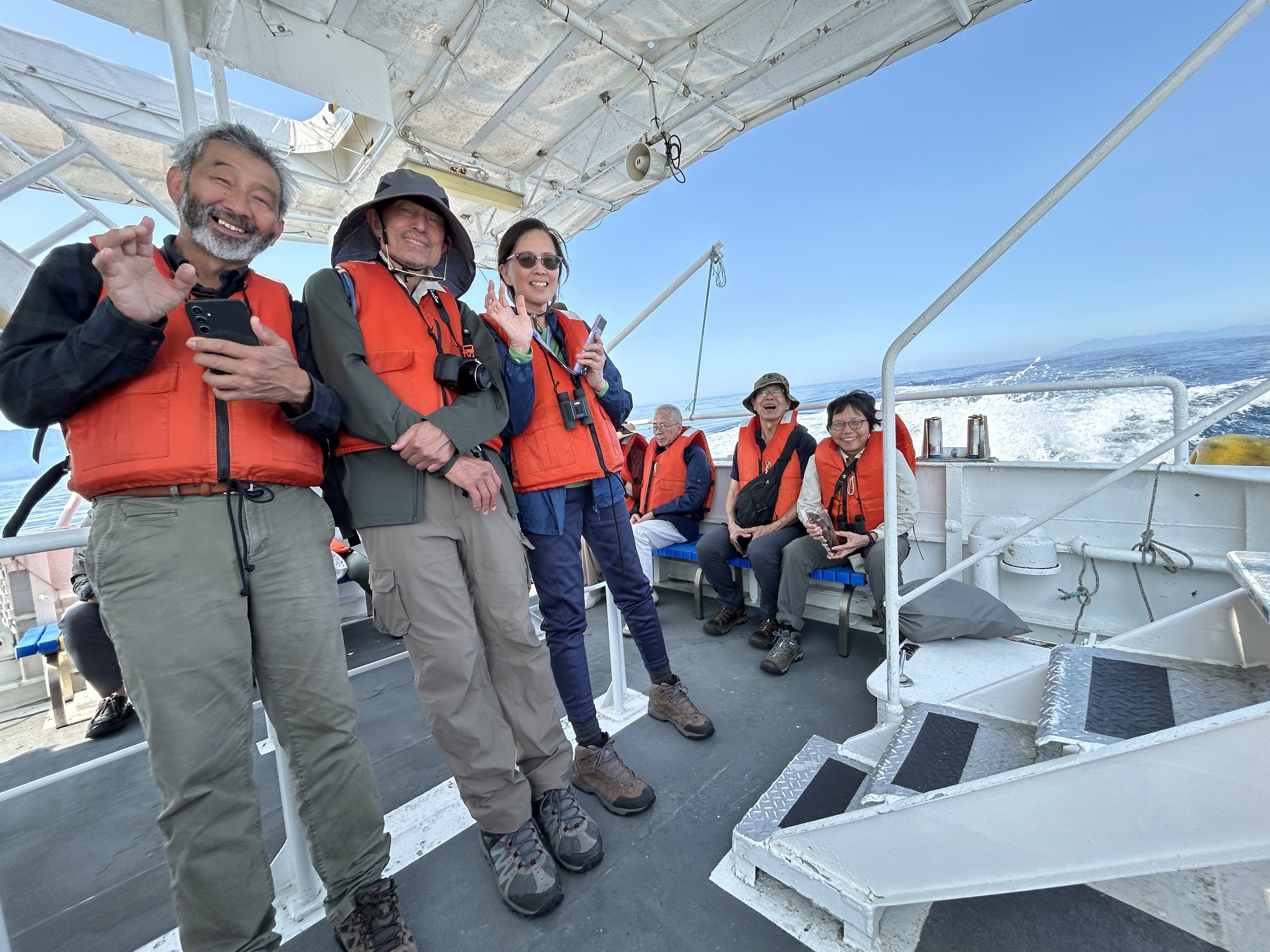 A group of people smile at the camera on board a boat. They are all wearing life jackets and are out at sea. It is a beautiful day and the sky is clear and blue.