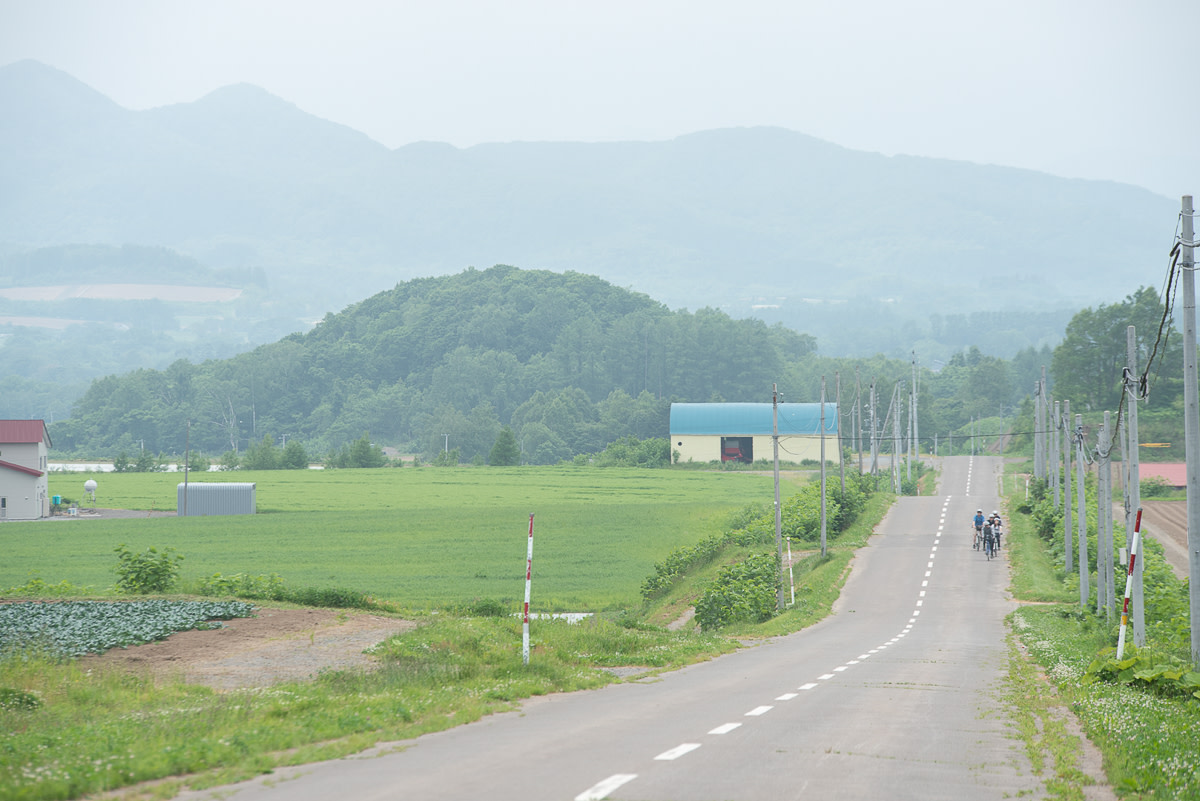 A group of cyclists are pictured in the distance riding along a quiet road. The road cuts through farm land is surrounded by fields. There is a barn and rolling hills in the distance.