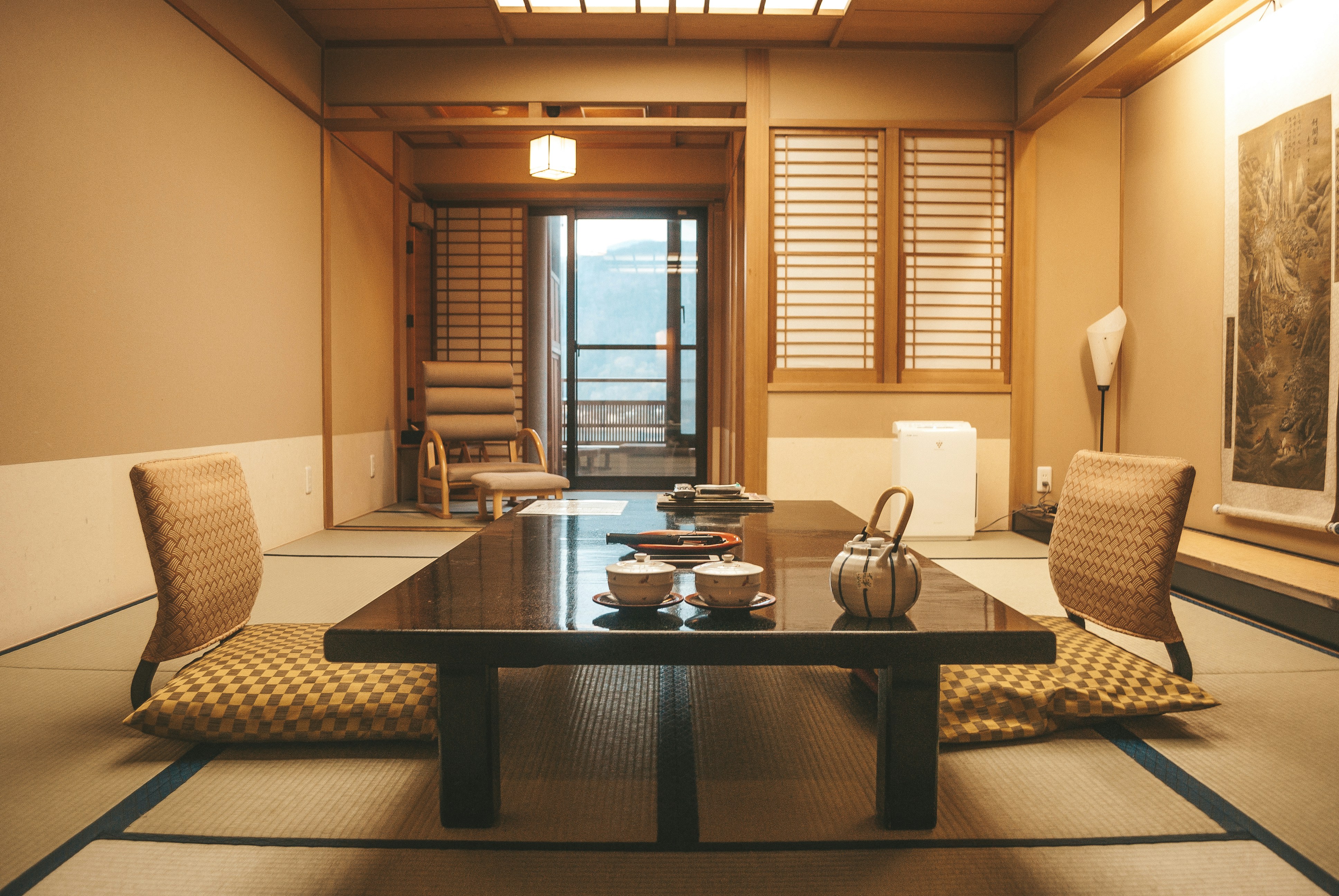 A Japanese bedroom with tatami mat flooring (made from dried rush grasses). There is a low table in the middle of the room with a Japanese tea set for two placed on it. At opposite ends of the table are two Japanese-style chairs (sat directly on the tatami with no legs).